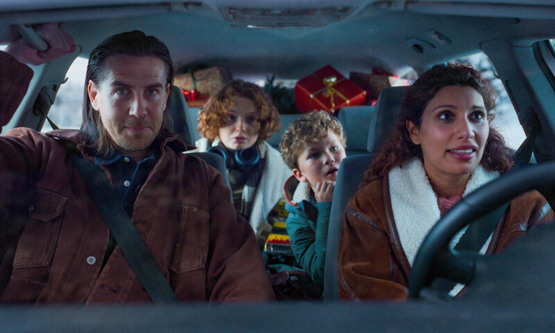 Familie im Auto, Erwachsene vorne, Kinder hinten. Geschenke im Fond. Es ist Tag, möglicherweise Winter.