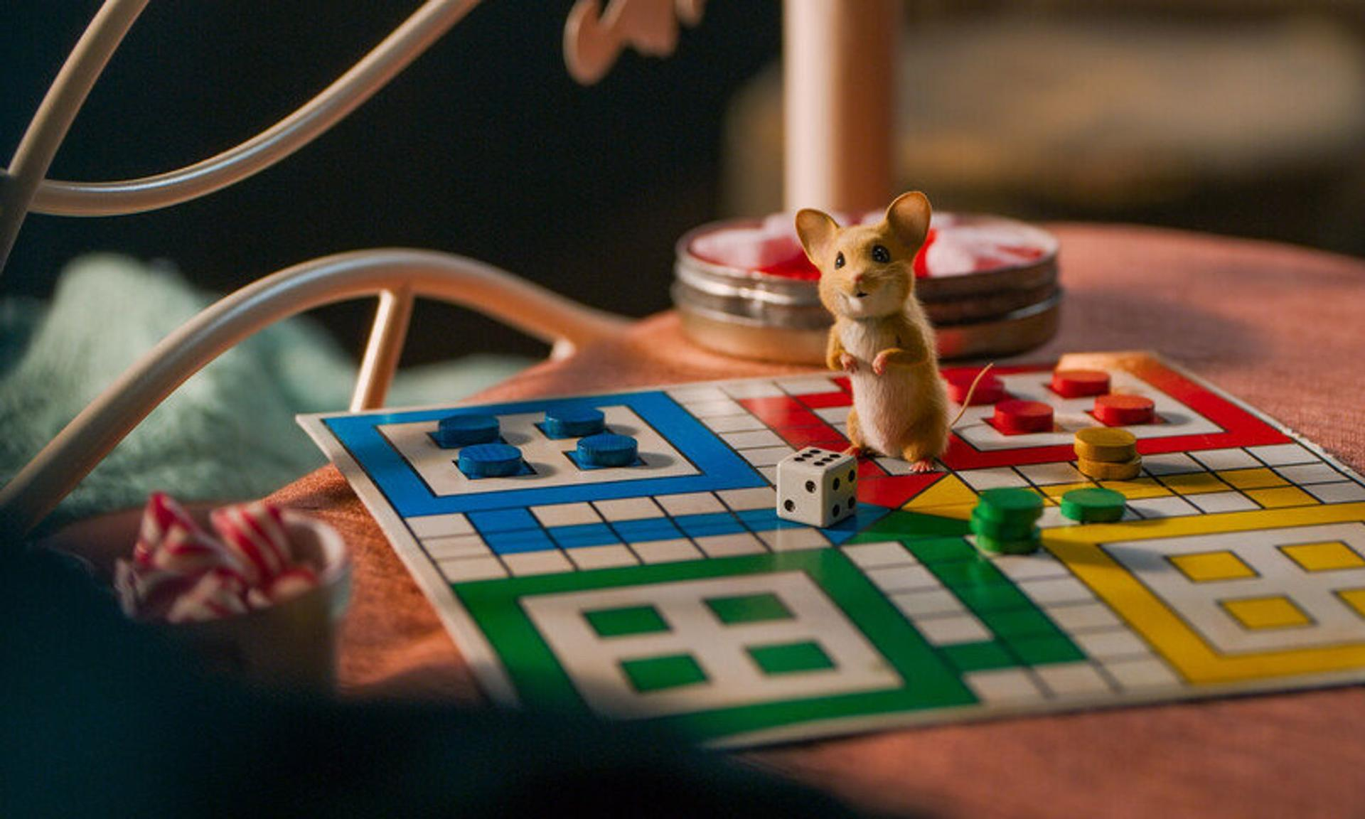 Eine Maus steht auf einem bunten Brettspiel. Daneben liegt ein Würfel. Rot, Grün, Gelb und Blau bilden das Spiel.