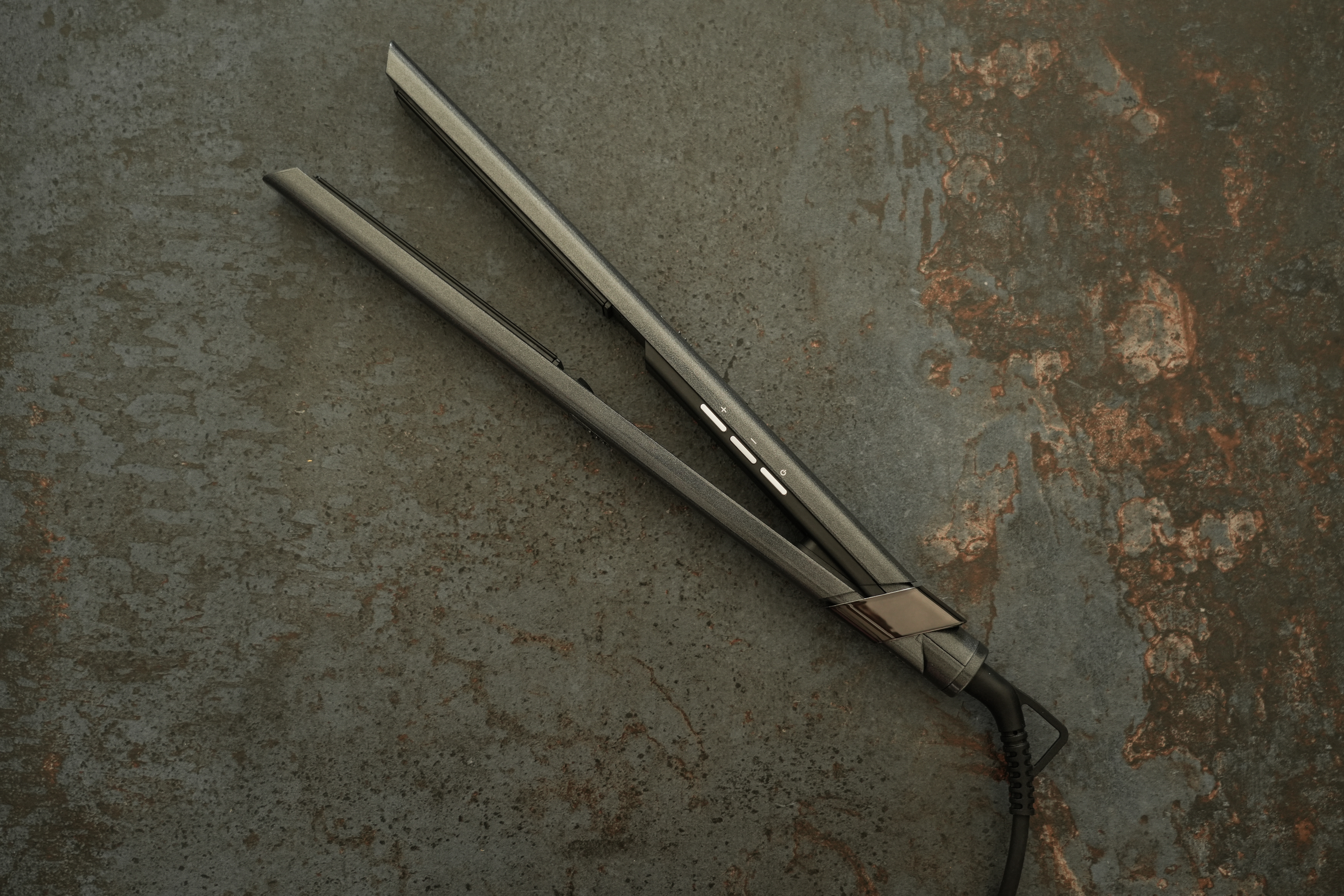 Prostownica na teksturowanej, ciemnoszarej powierzchni. Jest otwarta i pod kątem. Tło jest nakrapiane rdzawymi odcieniami.