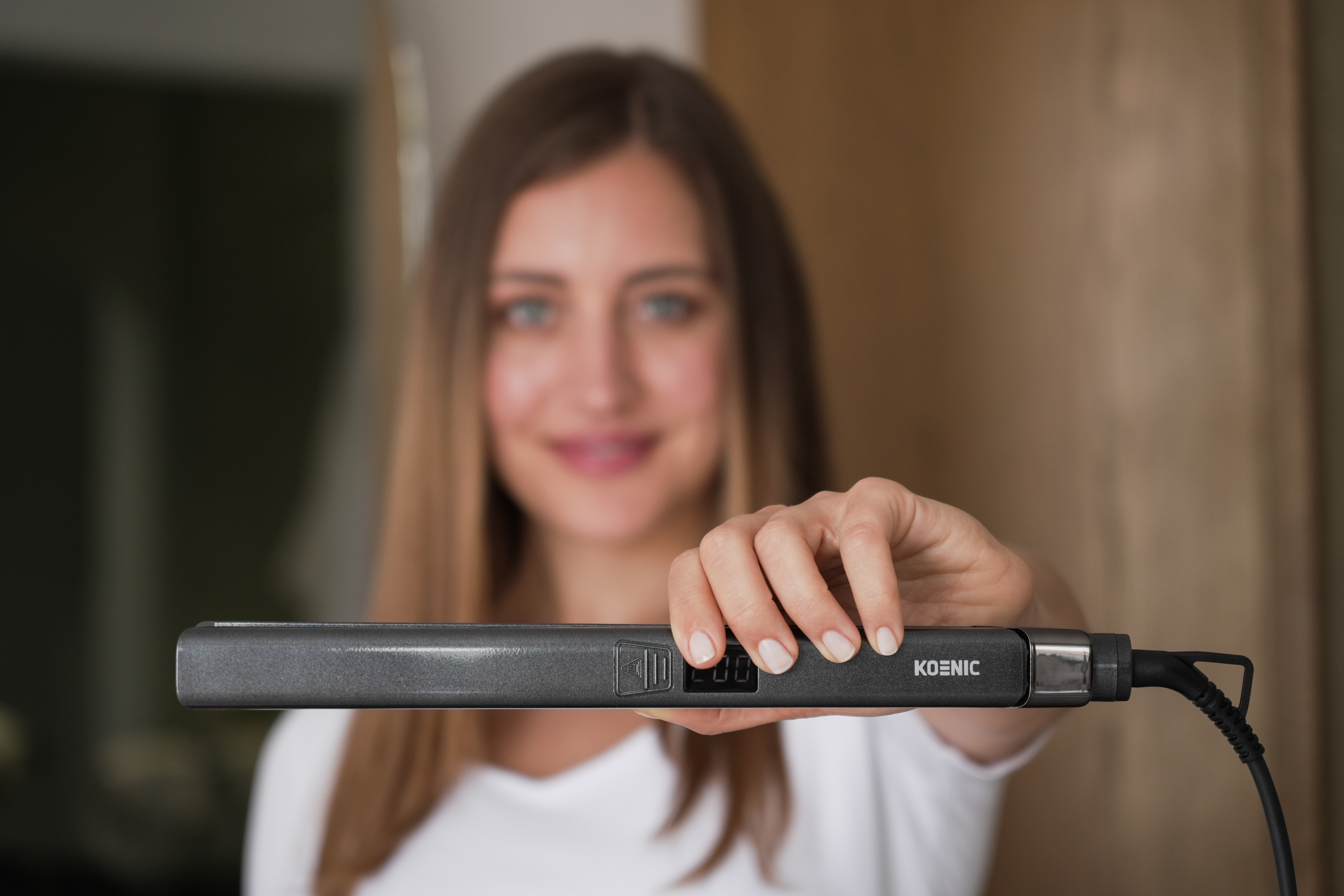 Kobieta trzyma prostownicę do włosów, twarz niewyraźna. Prostownica jest szara i ma markę Koenic.