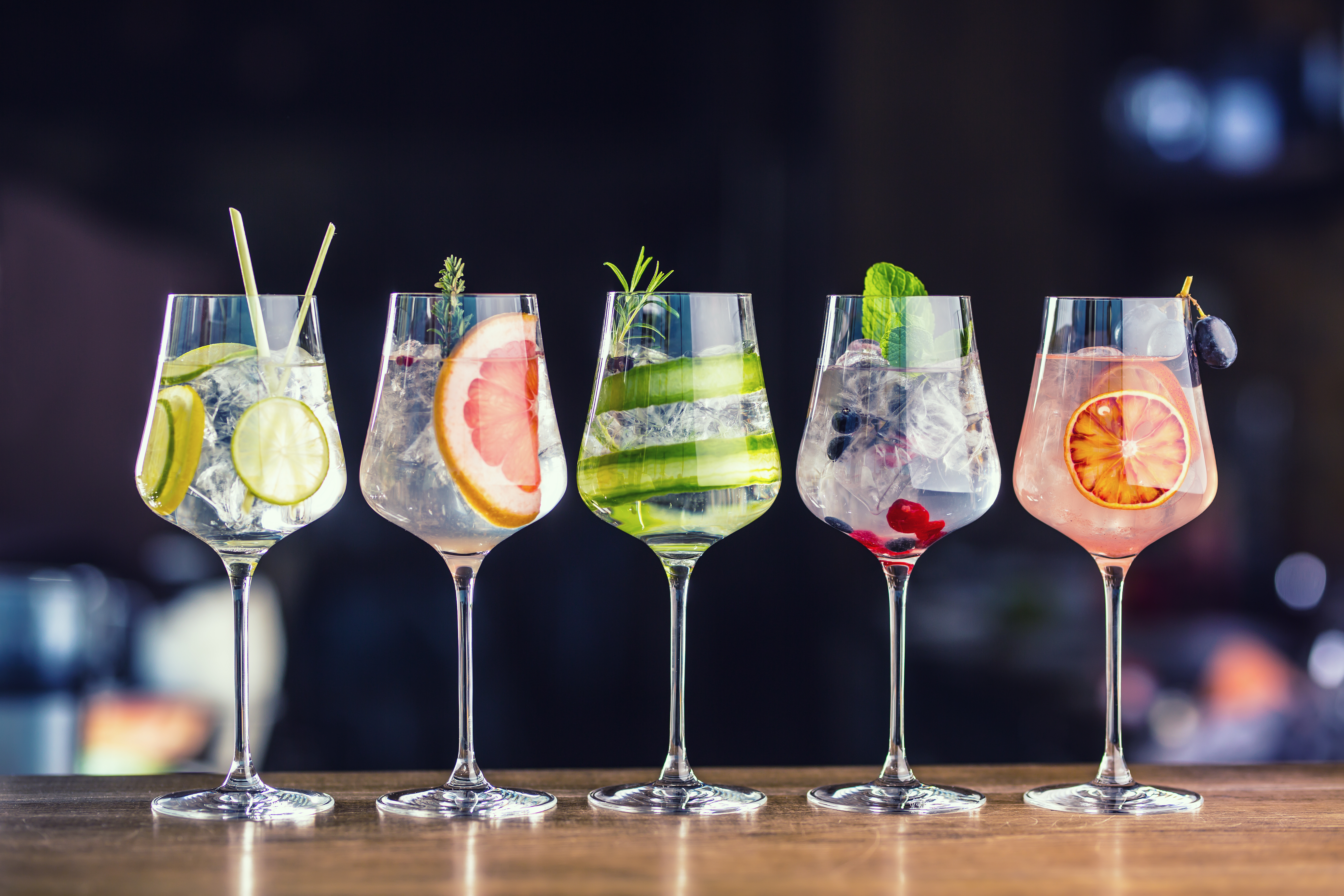 Pięć kieliszków do koktajli z różnymi dodatkami. Widoczne są limonki, grejpfruty, ogórki, mięta i plasterki pomarańczy.