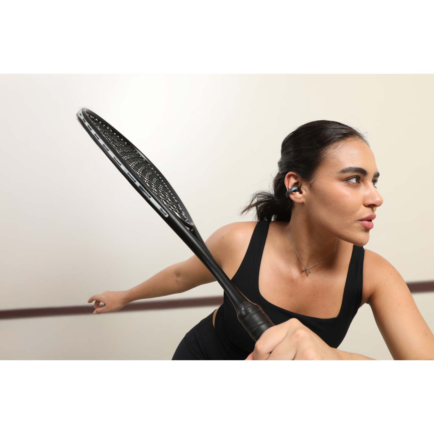 Une femme jouant au squash. Elle tient une raquette, porte une tenue noire et un écouteur. Le fond est un mur de couleur claire.