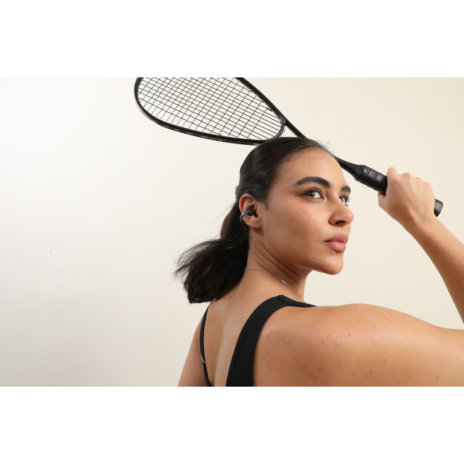 Une femme portant des écouteurs tient une raquette de tennis au-dessus de sa tête. Elle porte un haut noir et un collier argenté. Fond beige.