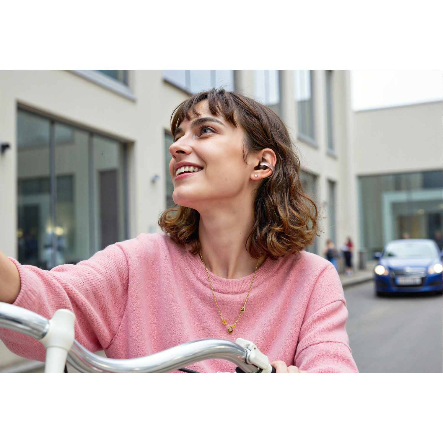 Une femme à vélo sourit, regardant vers le haut. Elle porte un pull rose et un collier. Des bâtiments et une voiture bleue en arrière-plan.