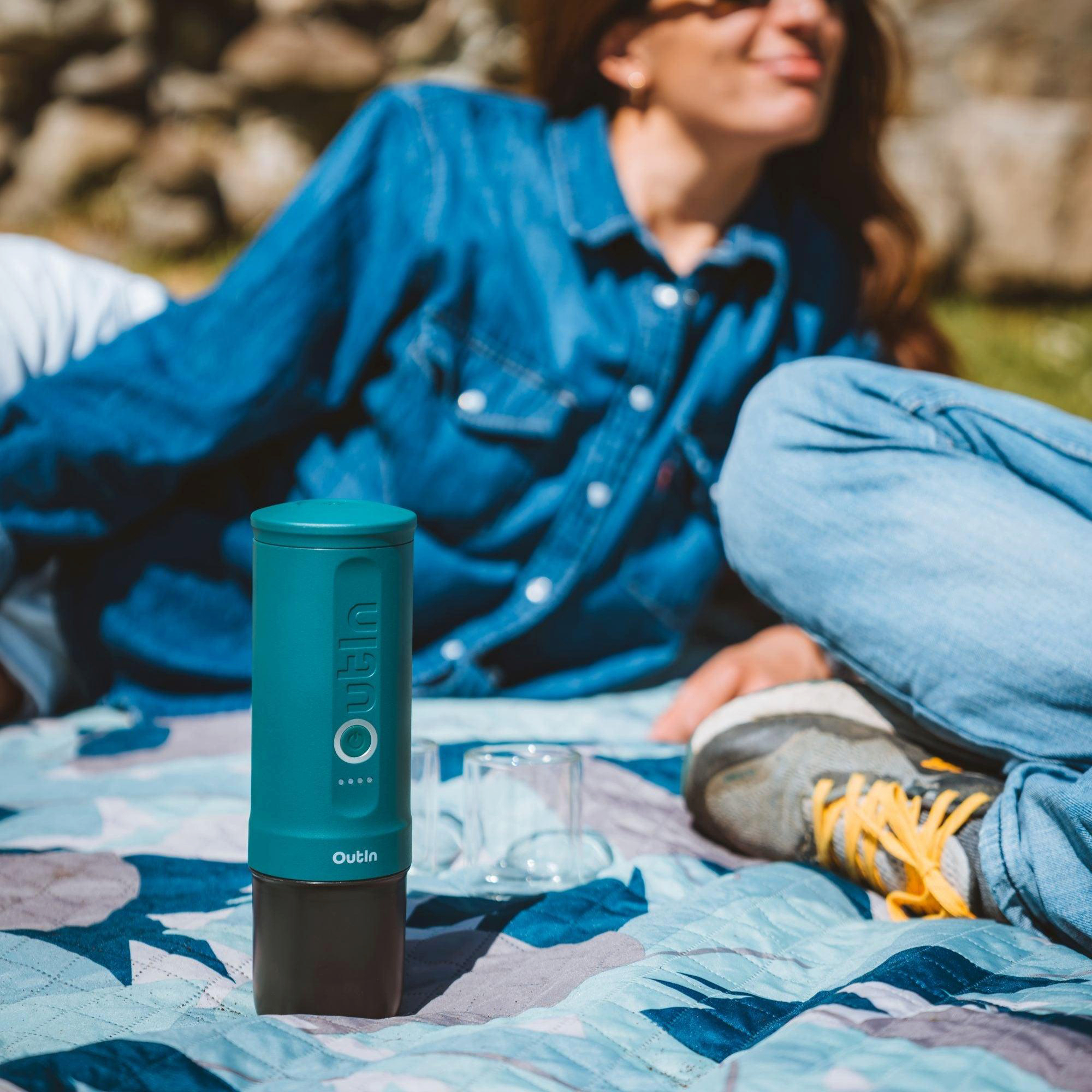 Eine Frau sitzt bei einem Picknick. Vorne steht ein türkisfarbener Kaffeezubereiter. Sie trägt Jeans und ein Jeanshemd. Ein sonniger Tag.