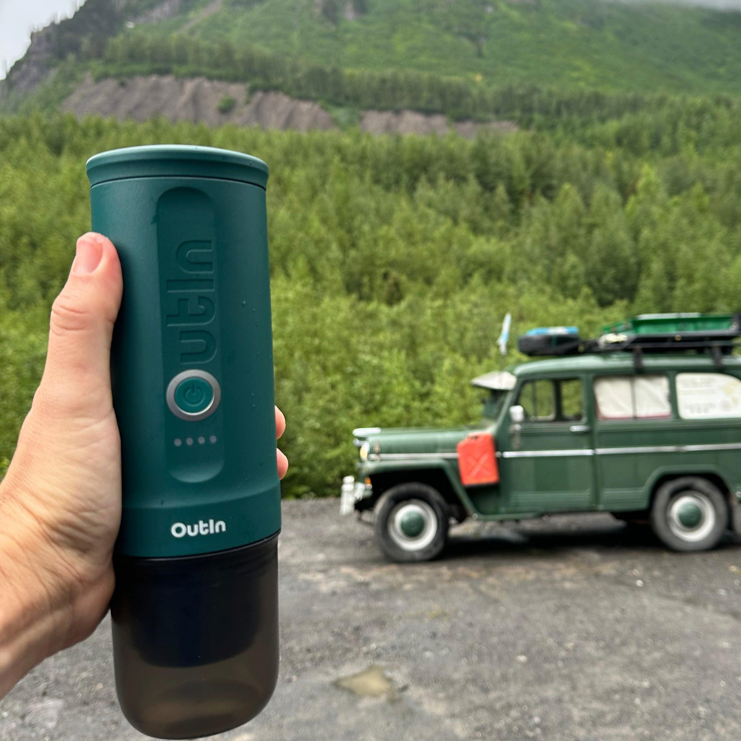 Eine Hand hält eine grüne Outin-Flasche. Im Hintergrund ein grüner Jeep. Berge und Bäume vervollständigen die Szene.