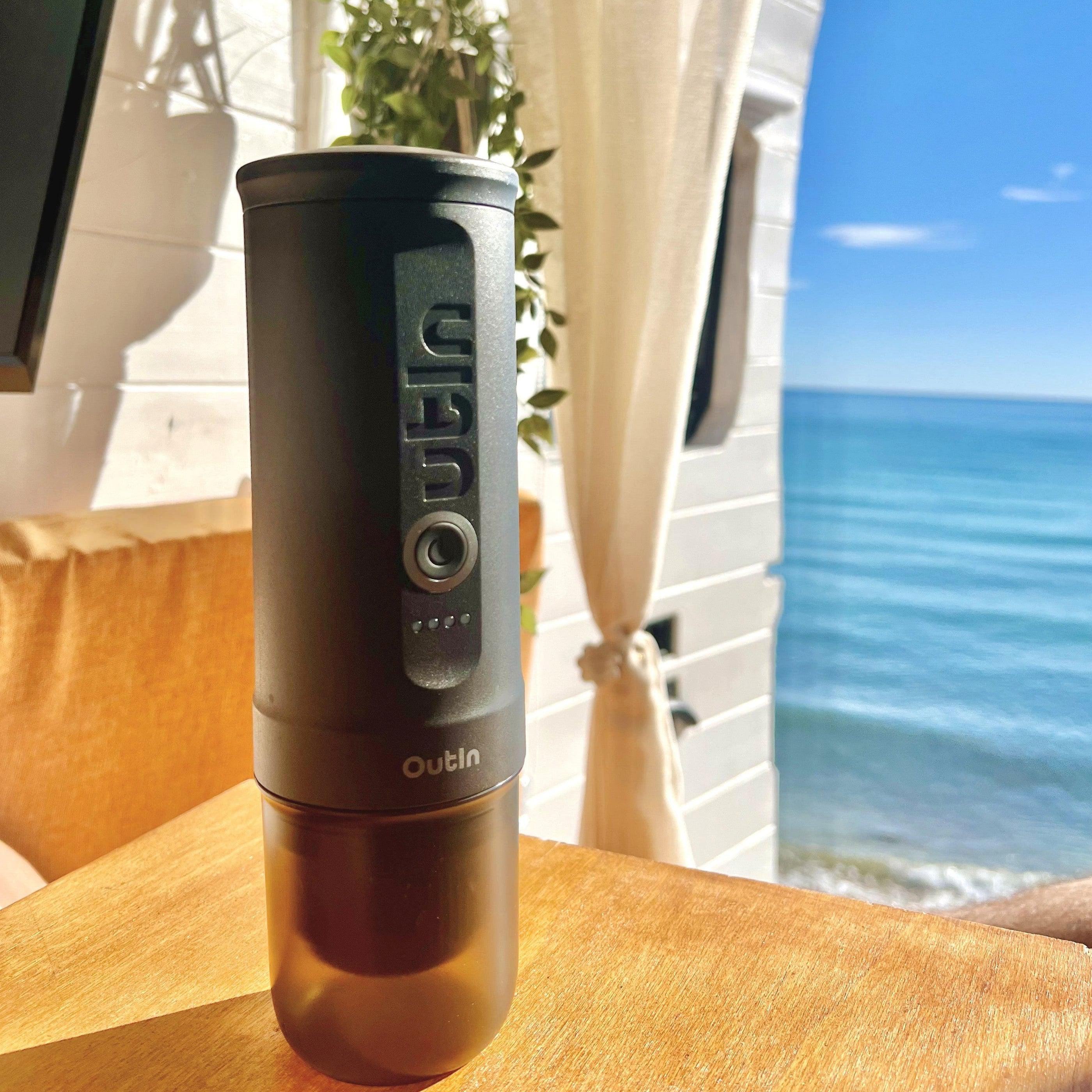 Ein schwarzer Outin-Kaffeebereiter steht auf einem Holztisch. Im Hintergrund sind das Meer und der blaue Himmel.