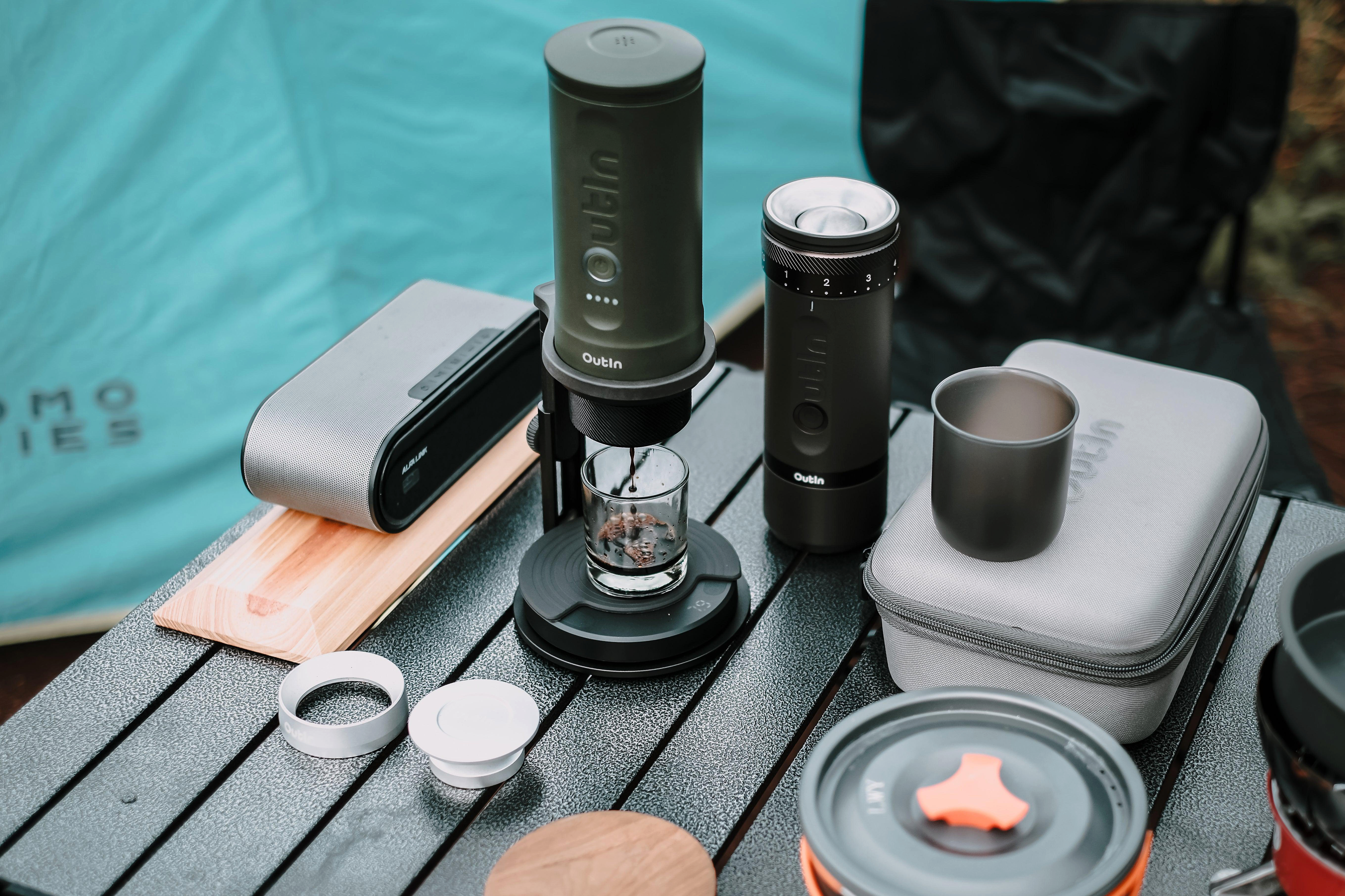 Kaffee-Setup auf einem Tisch. Enthält eine Mühle, ein Brühgerät, eine Tasse, eine Hülle und einen Lautsprecher, mit Zelt.
