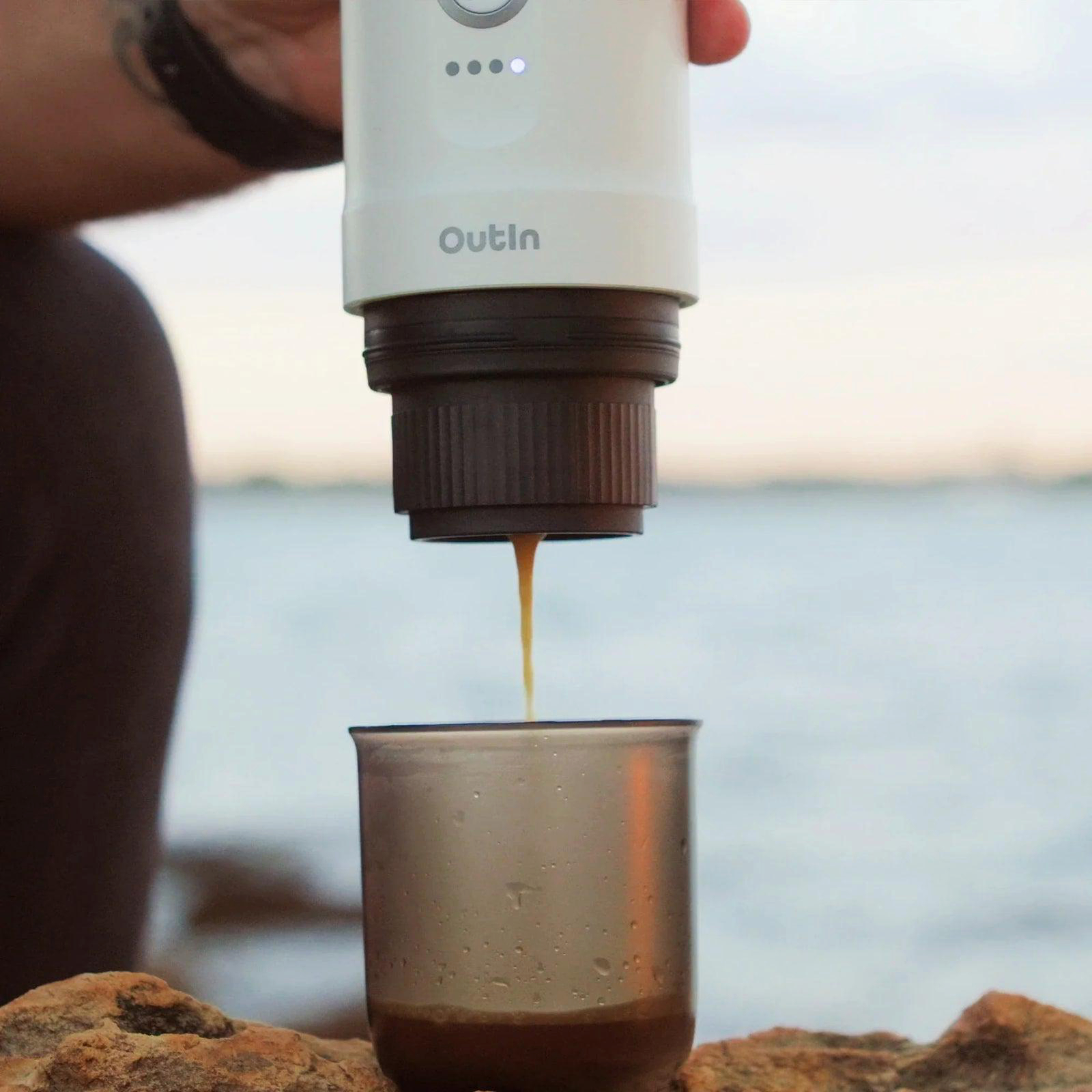 Kaffeemaschine, die Kaffee in ein Glas auf einem Felsen gießt, mit Wasser im Hintergrund.