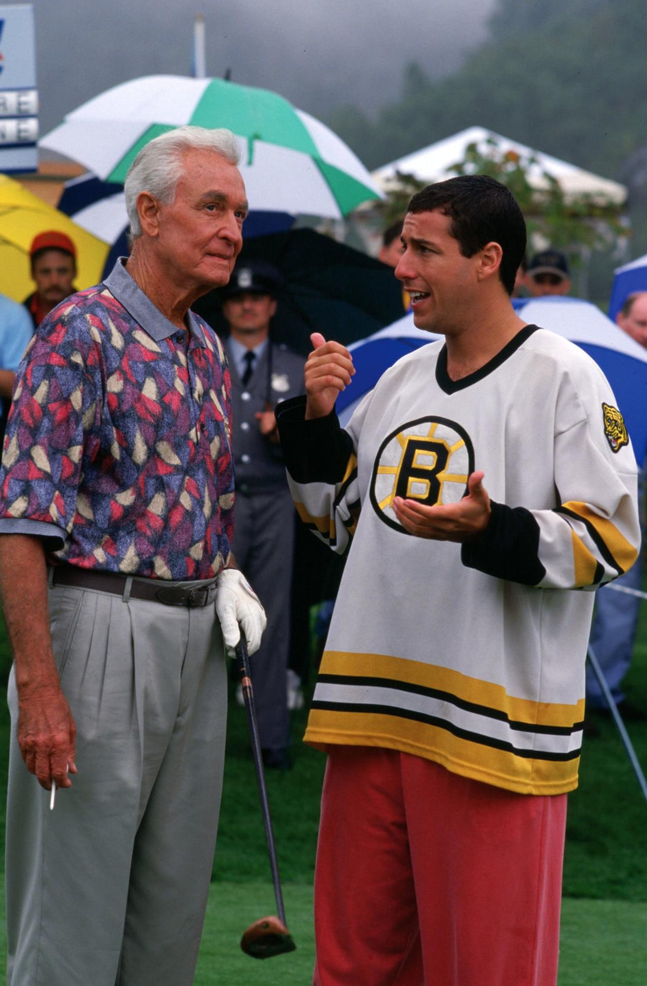 Zwei Männer auf einem Golfplatz, einer in einem gemusterten Hemd, der andere in einem Bruins-Trikot. Im Hintergrund sind Schirme und Leute zu sehen.