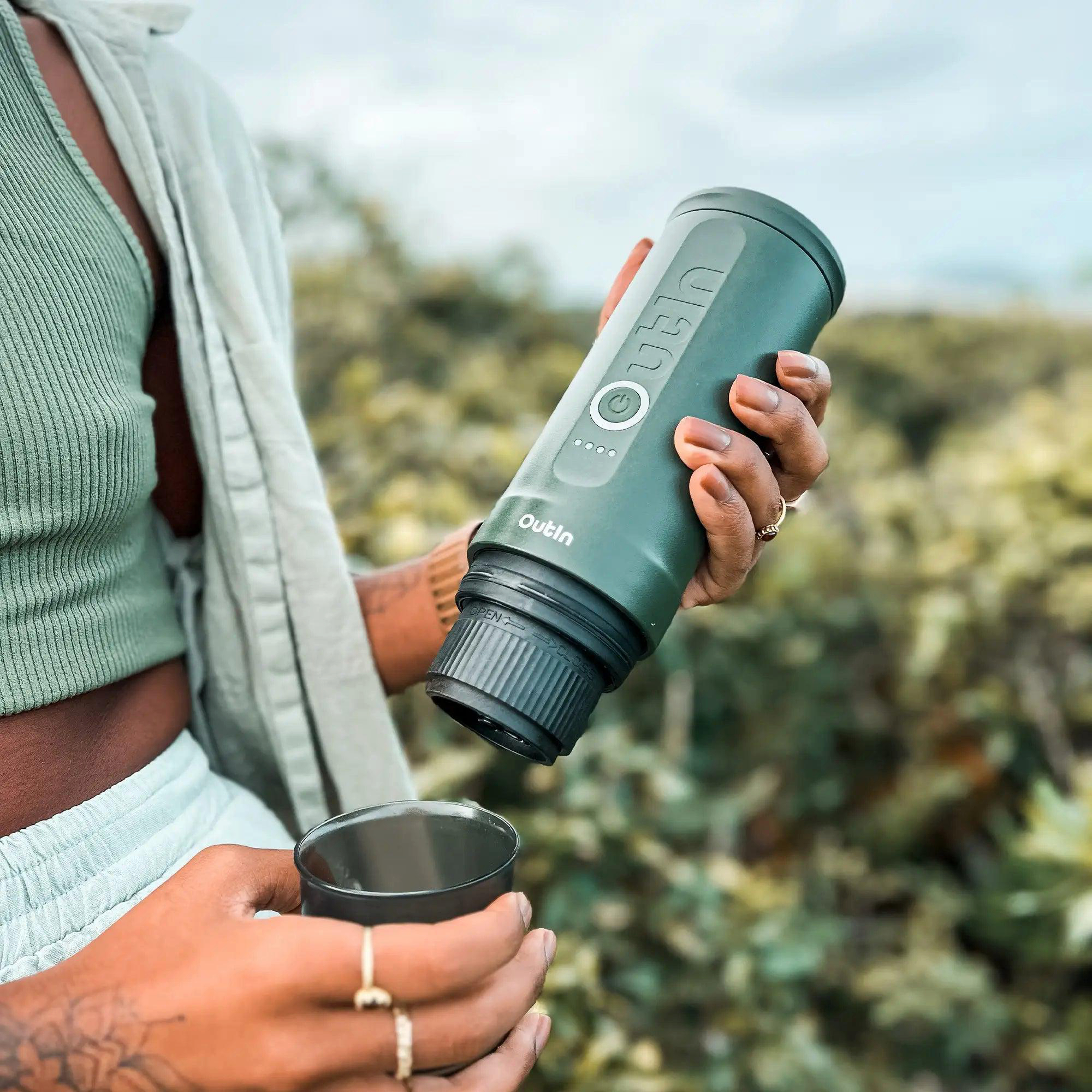 Eine Person hält draußen einen grünen Wasserfilter und eine schwarze Tasse. Der Filter ist mit 'Outin' beschriftet.