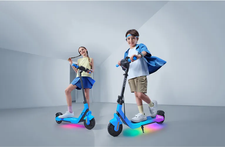 Két gyerek elektromos robogón. Az egyik lány a copfjait fogja, a fiú mosolyog. Mindkét robogón színes lámpák vannak.