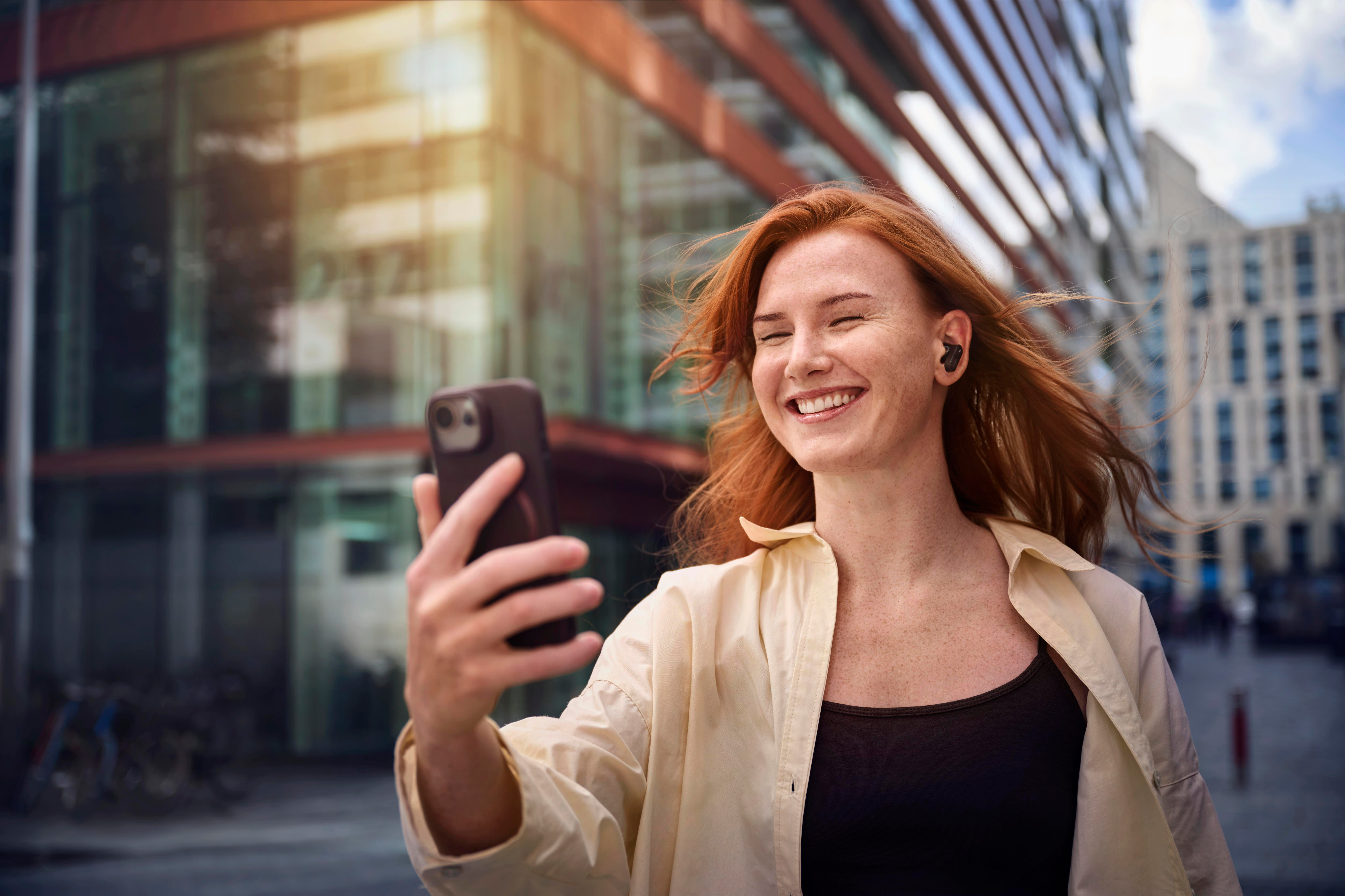 Eine rothaarige Frau lächelt und hält ein Telefon. Sie trägt ein schwarzes Hemd und eine beige Jacke. Gebäude im Hintergrund.