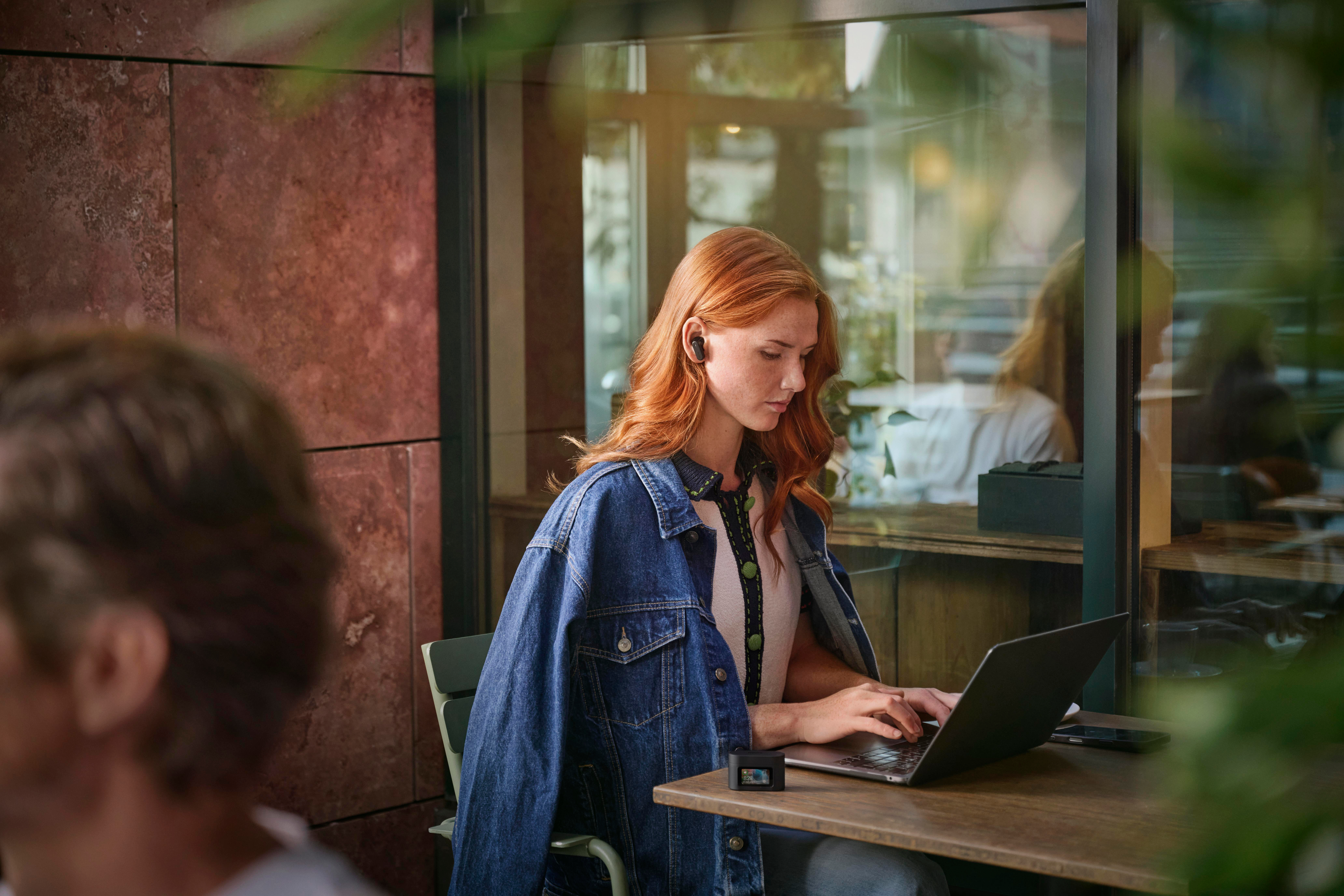 Eine rothaarige Frau mit Jeansjacke benutzt einen Laptop an einem Cafétisch. Sie hat Ohrhörer im Ohr. Der Hintergrund ist unscharf.