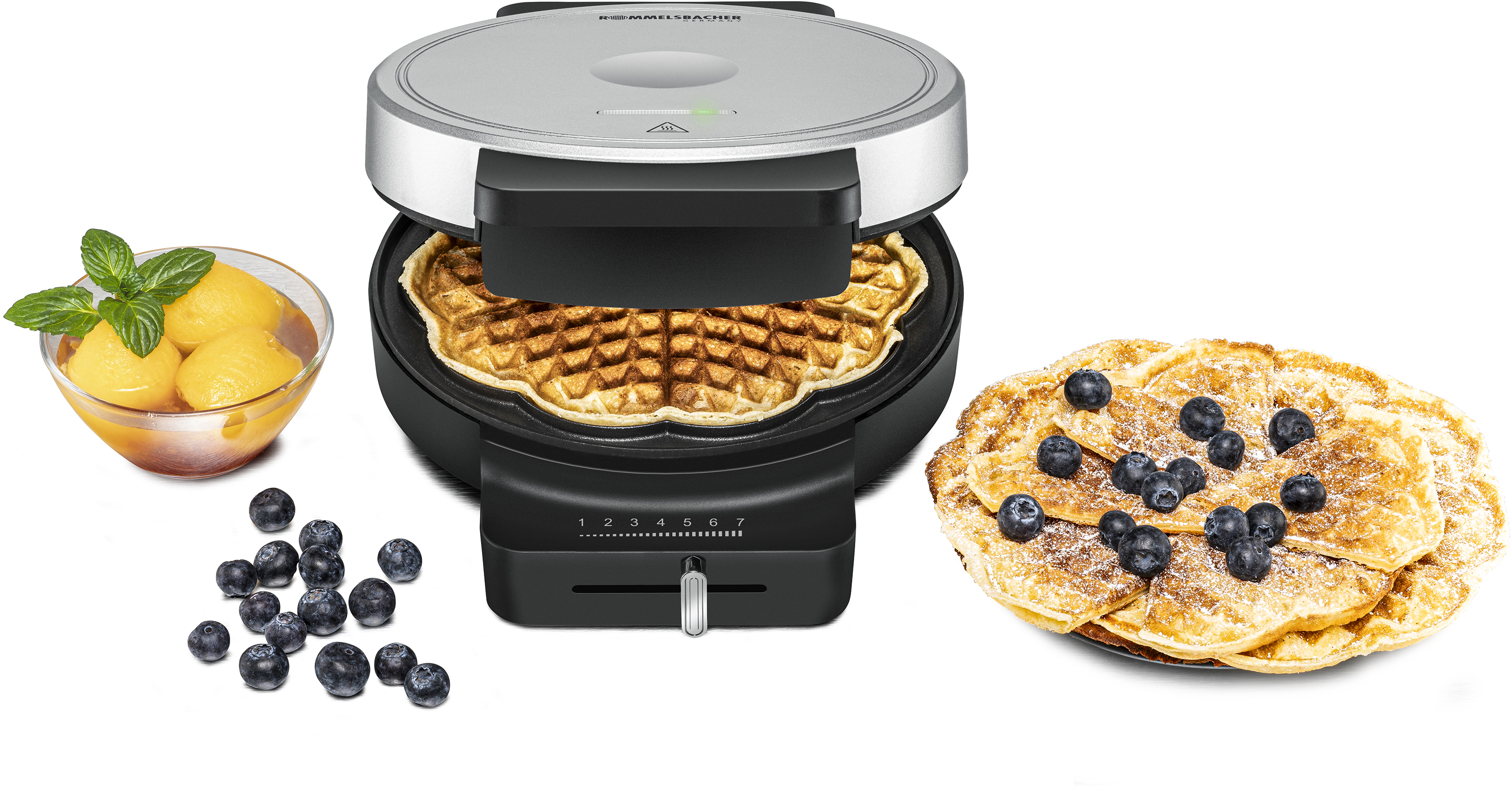 Waffeleisen mit fertiger Waffel. Daneben Blaubeeren und eine Schüssel Pfirsiche. Außerdem ein Teller Waffeln mit Blaubeeren.