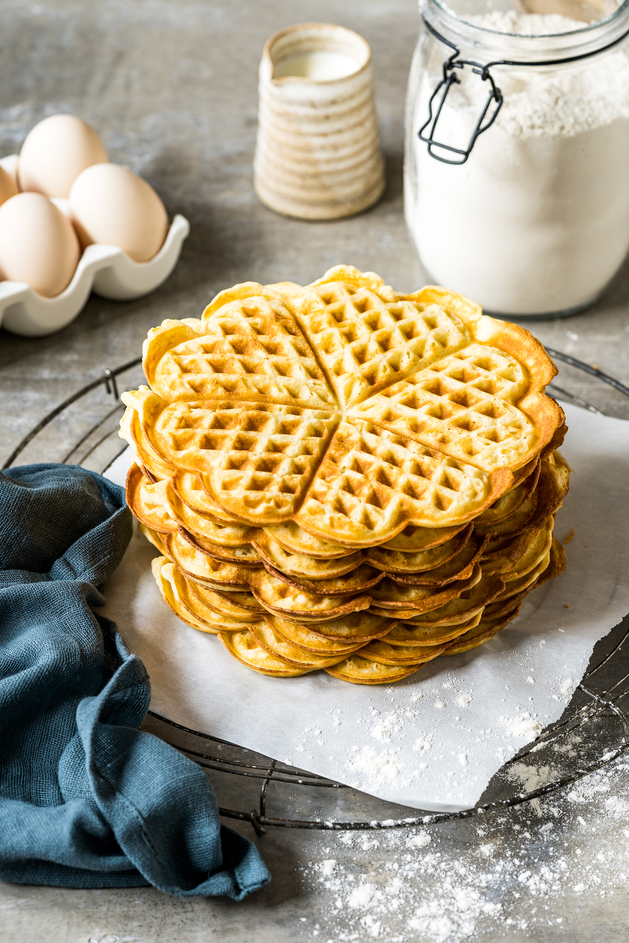 Stapel goldener Waffeln auf Pergamentpapier. Eier, Mehl und Milch im Hintergrund. Links ein blaues Tuch.