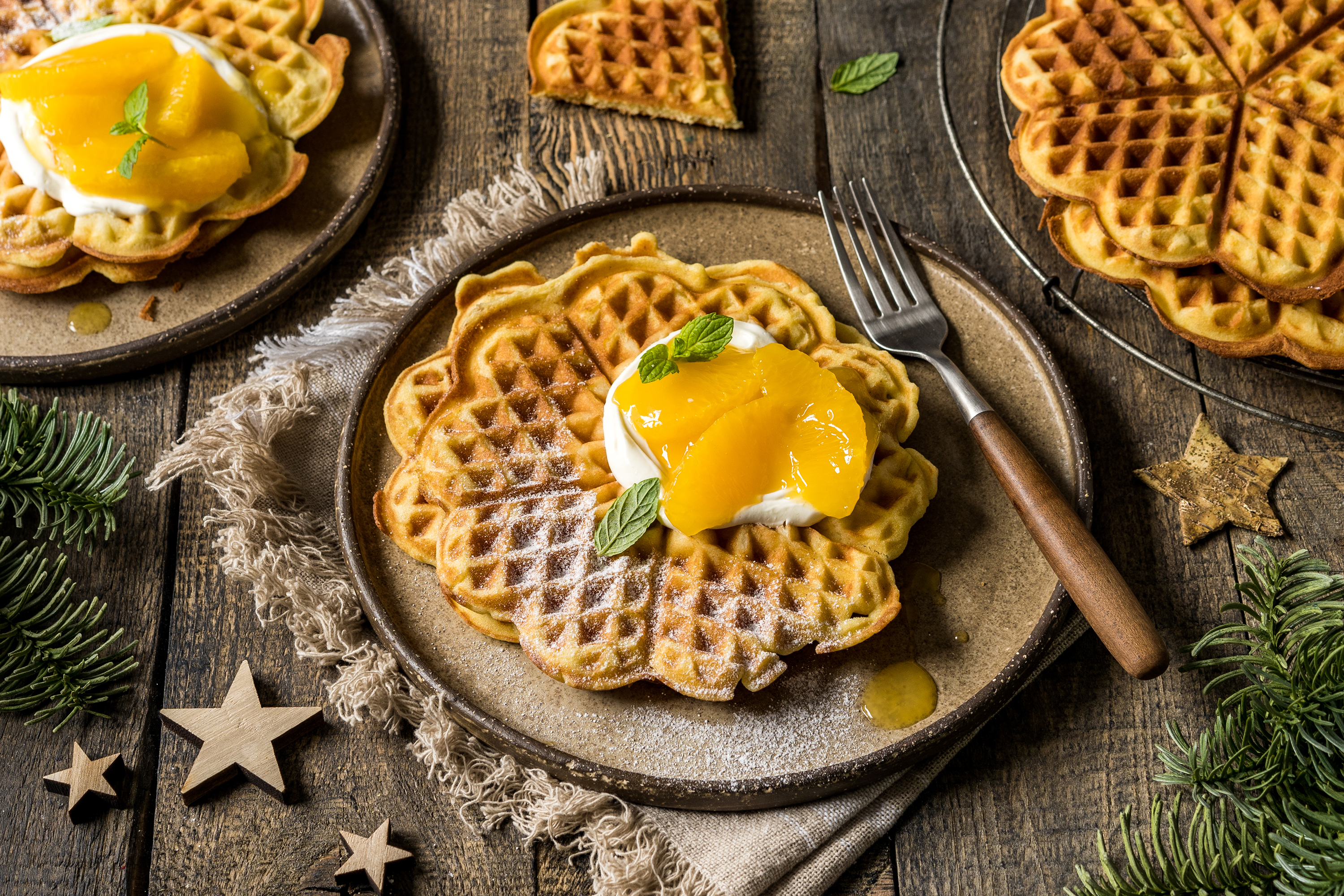 Waffeln auf Tellern, belegt mit Sahne und Pfirsichen, Minzblättern. Holztisch mit Gabel, Stern-Deko und Grün.