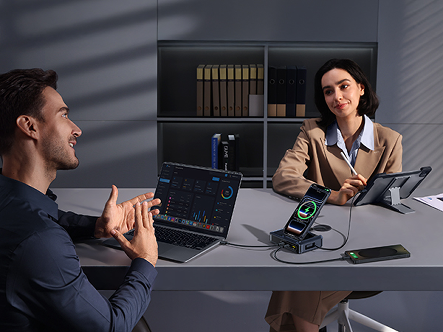 Dos personas en un escritorio con electrónica. Se ven un portátil, una tableta y teléfonos. Un teléfono se está cargando. Ambas personas sonríen.