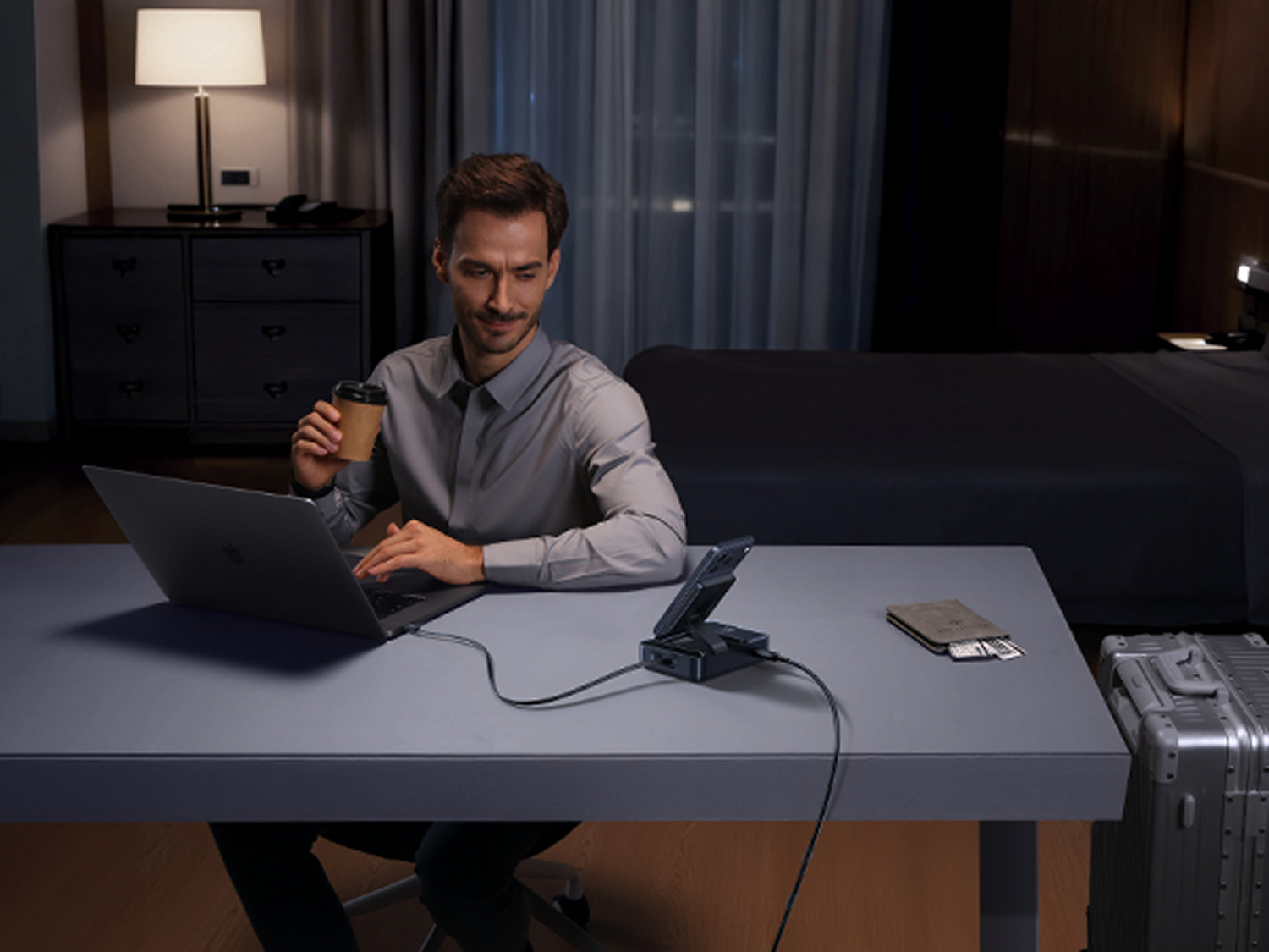 Un hombre con camisa está sentado en un escritorio, usando una laptop, sosteniendo una taza de café. Un cargador, una cartera y una maleta están en el escritorio.
