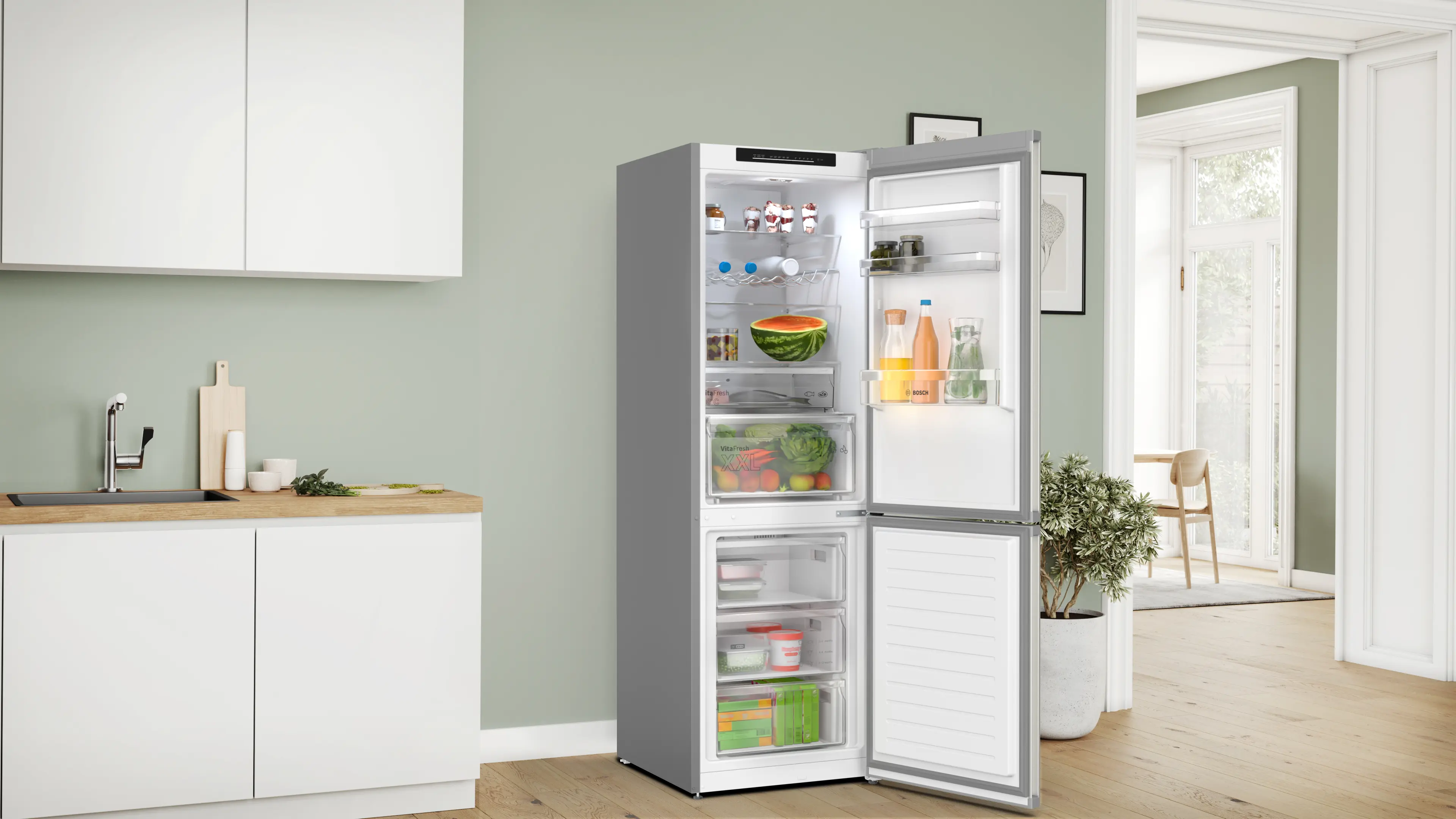 Refrigerador abierto con frutas y verduras. Cocina con gabinetes blancos y una planta. Pared verde claro y puerta abierta al exterior.