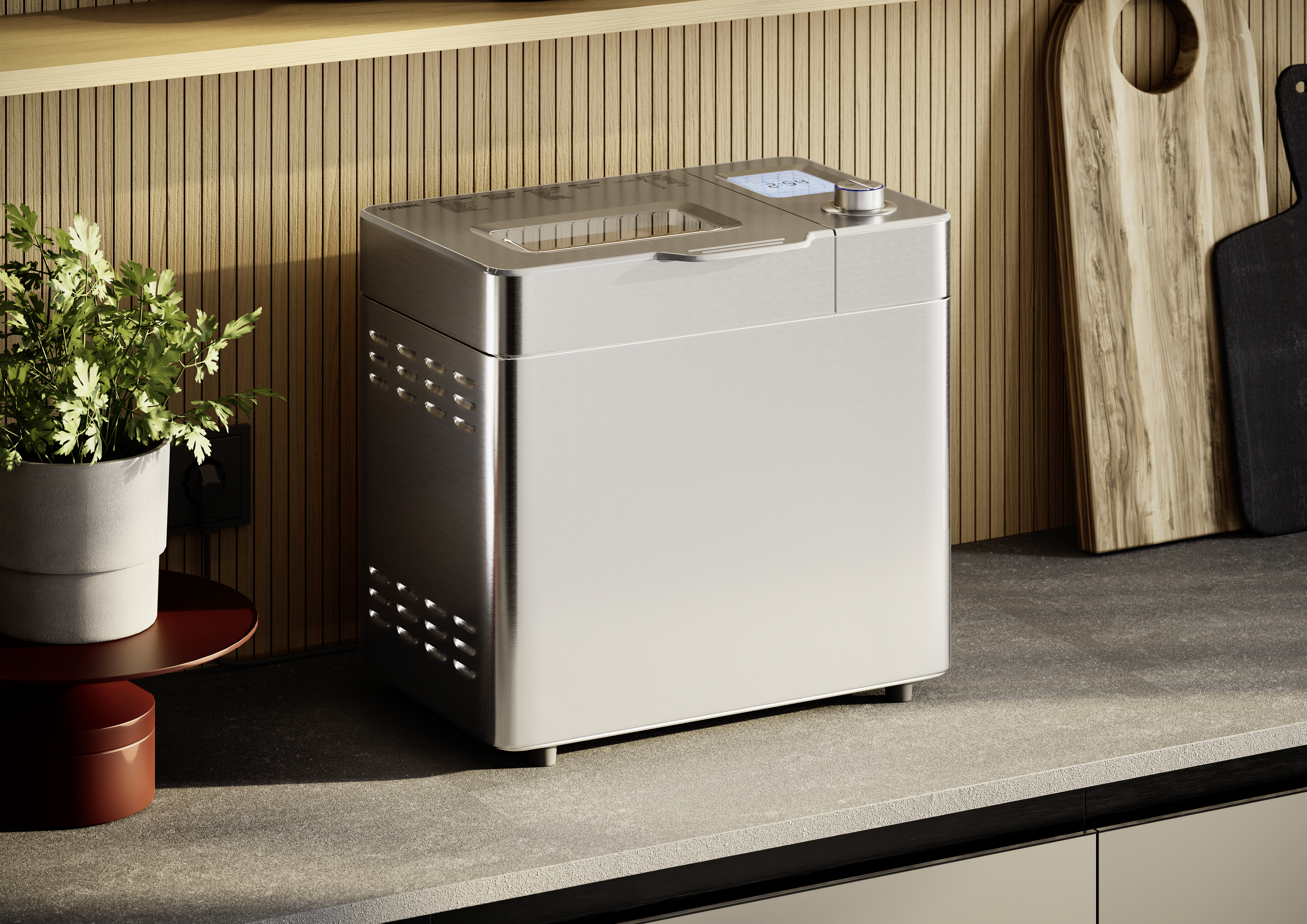 Ein Brotbackautomat aus Edelstahl auf einer Arbeitsplatte, mit einer Pflanze, Schneidebrettern und Holzverkleidung im Hintergrund.