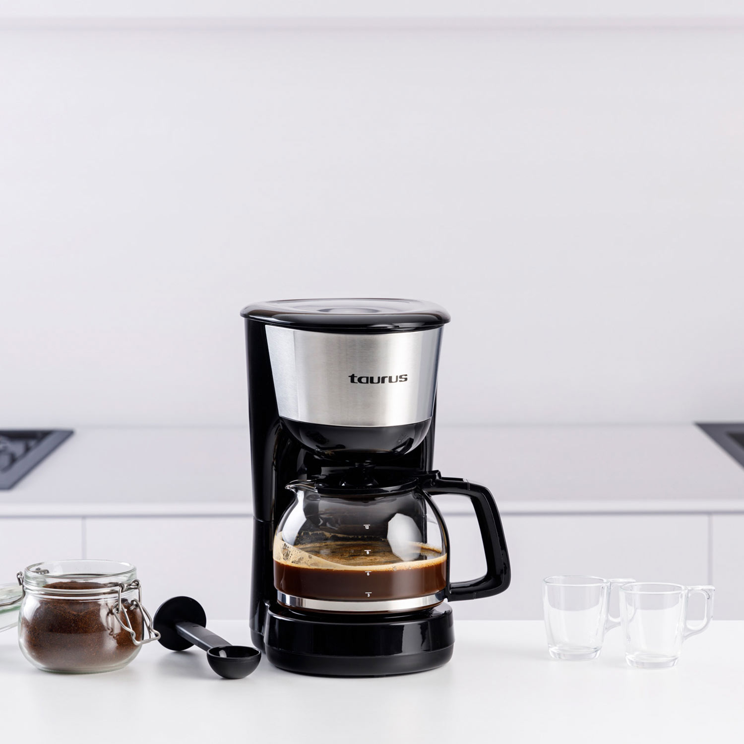 Cafetera con jarra de vidrio llena de café, junto a dos tazas de vidrio vacías, café molido y una cuchara en una superficie blanca.
