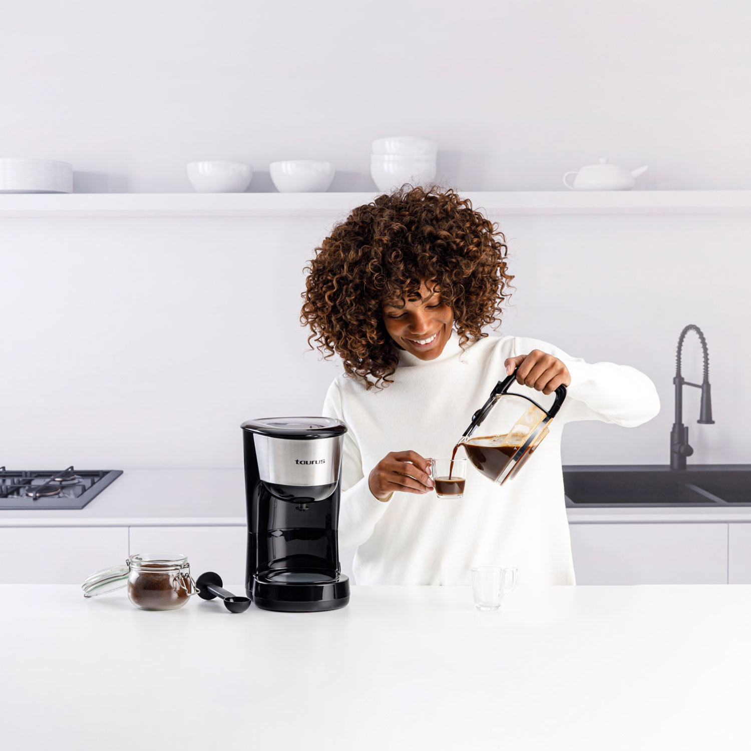 Una mujer vierte café en un vaso. En la encimera hay una cafetera, un tarro de café y una cuchara. Fondo de cocina blanco.
