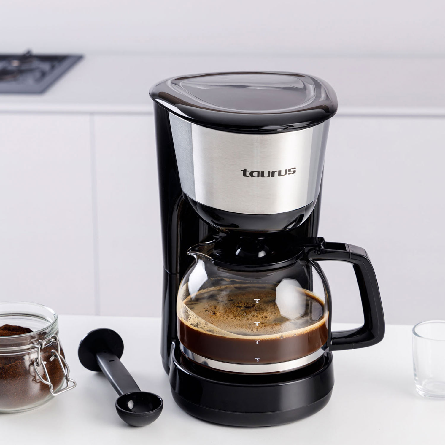 Una cafetera Taurus negra y plateada con una jarra de vidrio llena de café. Café molido y una cuchara están sobre la mesa.