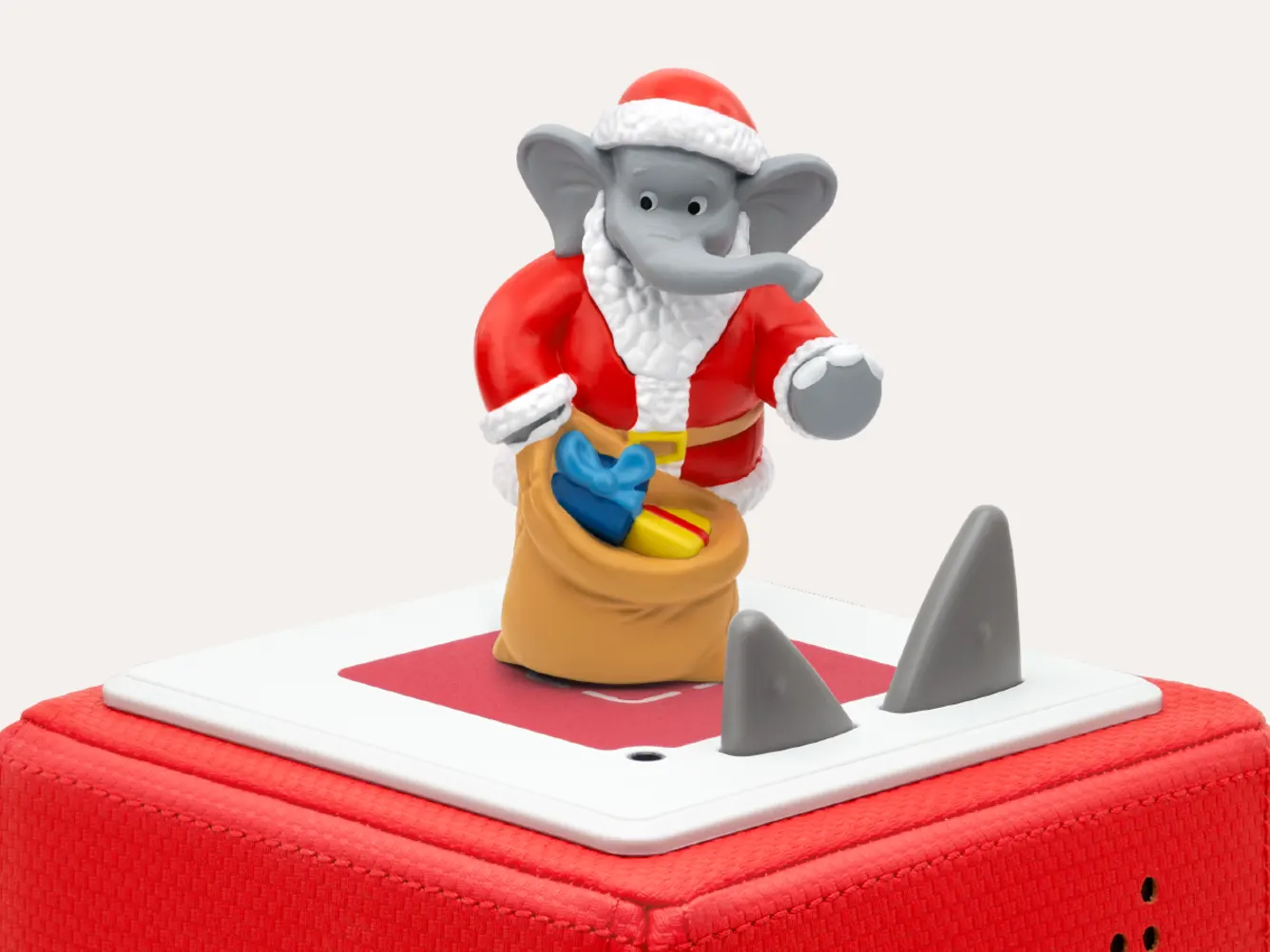 Un elefante vestito da Babbo Natale tiene in mano un sacco di regali. La figura è su una scatola rossa con forme grigie a forma di pinna di squalo.
