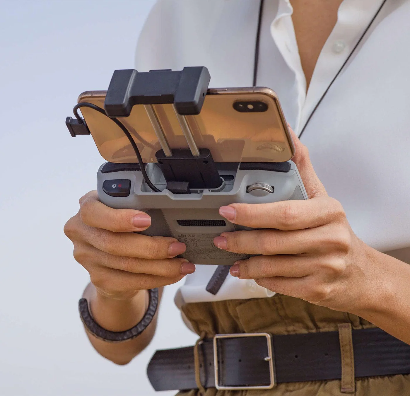 Una persona tiene in mano un controller per droni con un telefono collegato. Indossa una camicia bianca e una cintura marrone.