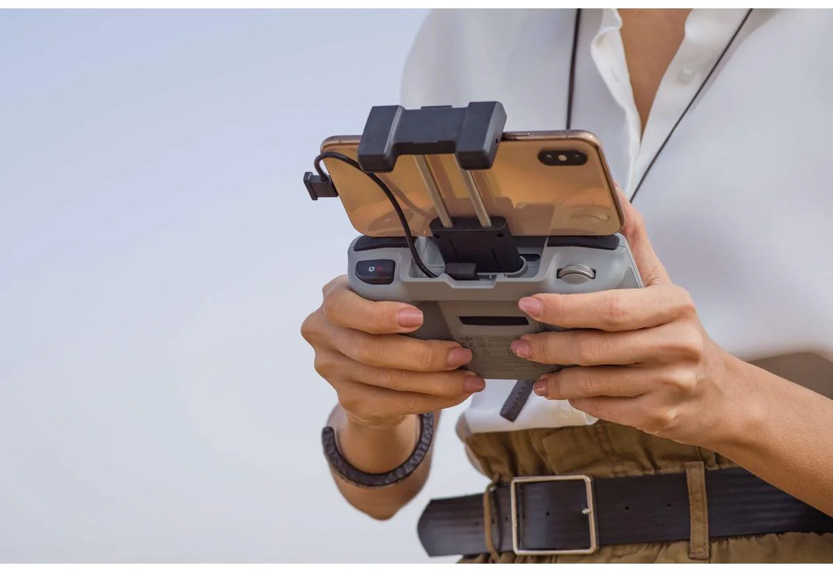 Eine Person hält eine Drohnenfernbedienung mit einem angeschlossenen Telefon. Sie trägt ein weißes Hemd und khakifarbene Hose mit schwarzem Gürtel.