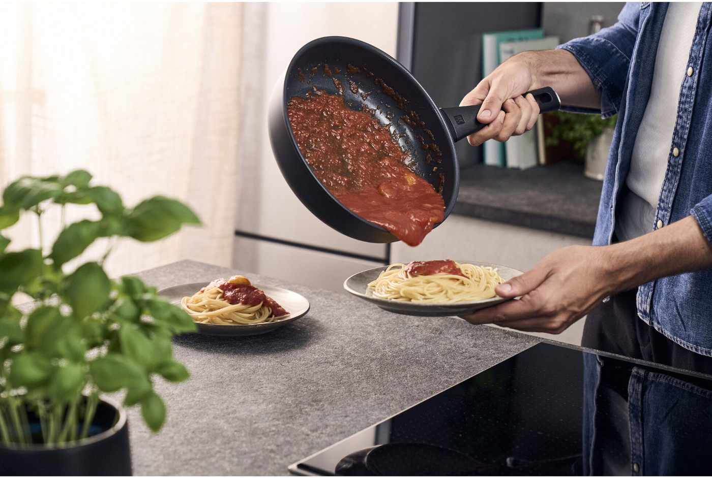 Eine Person gießt Tomatensauce aus einer Pfanne auf einen Teller Spaghetti. Ein weiterer Teller Spaghetti befindet sich in der Nähe. Grüne Pflanze im Vordergrund.