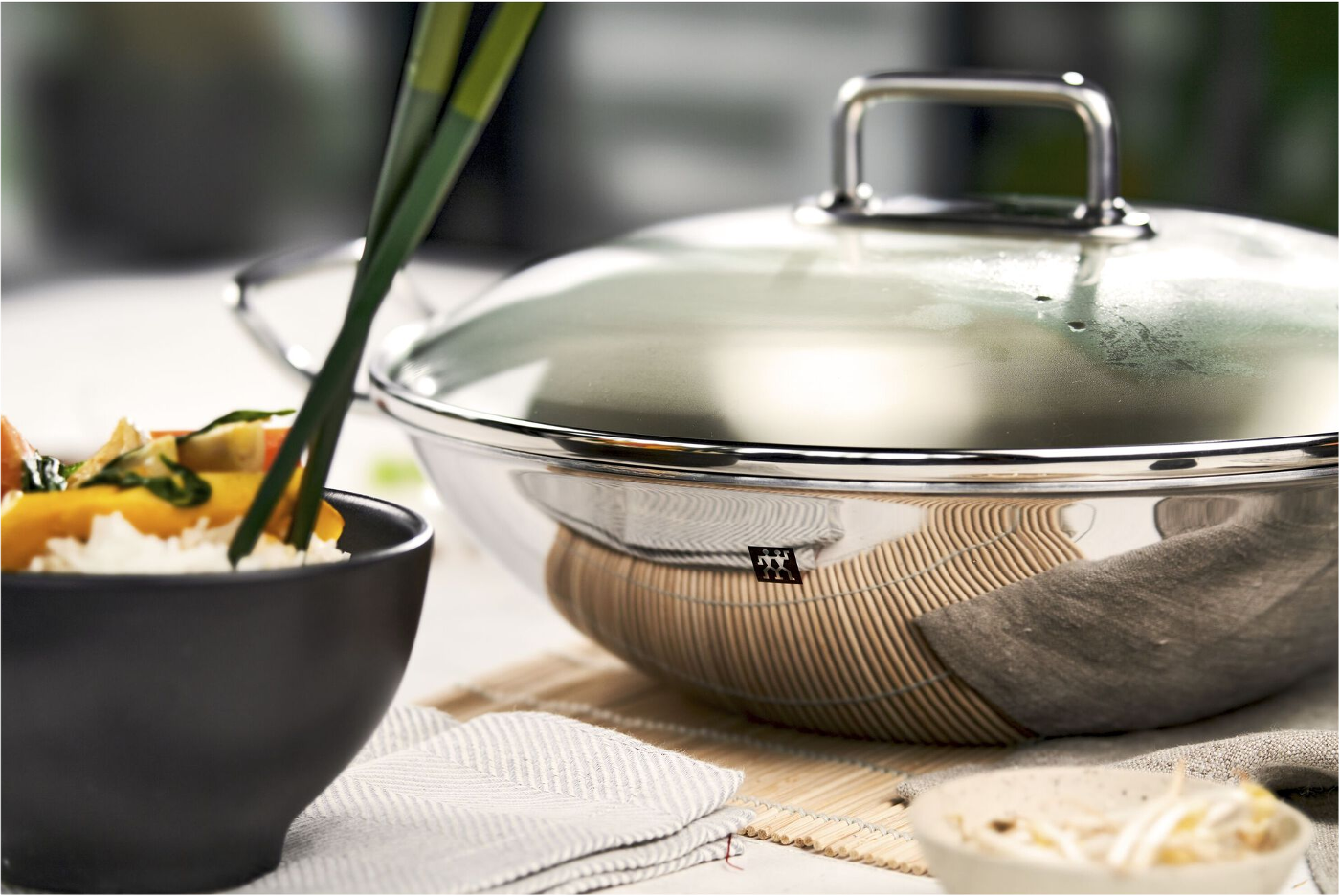 Eine Edelstahl-Wok mit Glasdeckel, Essstäbchen und eine Schüssel Reis mit Gemüse stehen auf einem Tisch.