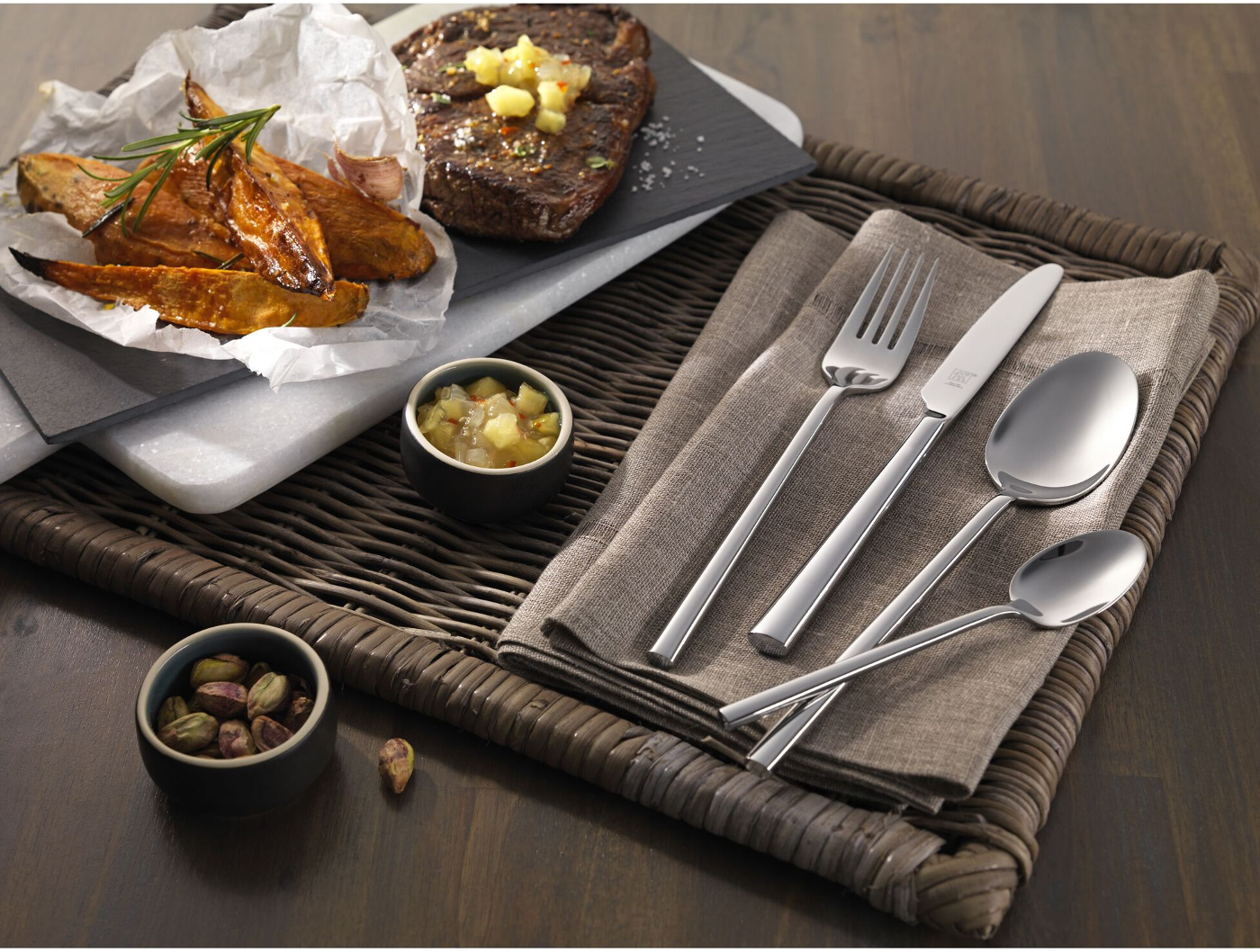 Ein Tischarrangement mit Steak, Süßkartoffelpommes und Besteck auf einem geflochtenen Tablett. Pistazien und Chutney sind ebenfalls vorhanden.