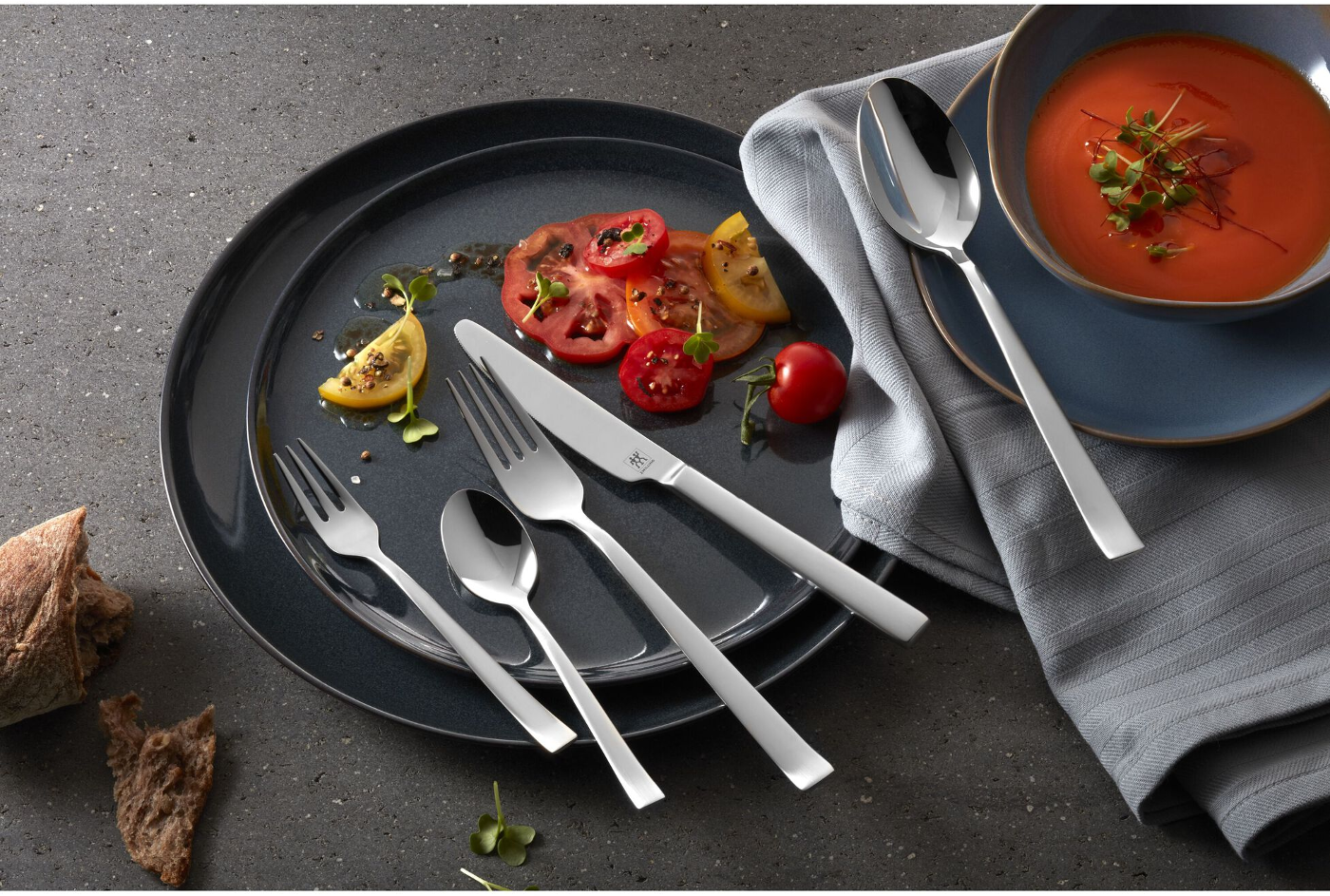 Ein Gedeck mit Tomatenscheiben, Besteck, einer Schüssel Suppe und einem Stück Brot auf dunkler grauer Oberfläche.