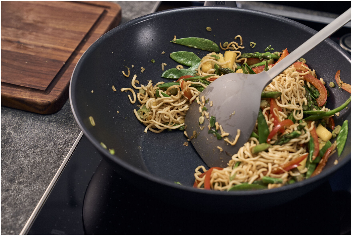 Ein Wok mit Nudeln, Gemüse und einem Spatel. Ein hölzernes Schneidebrett befindet sich im Hintergrund.
