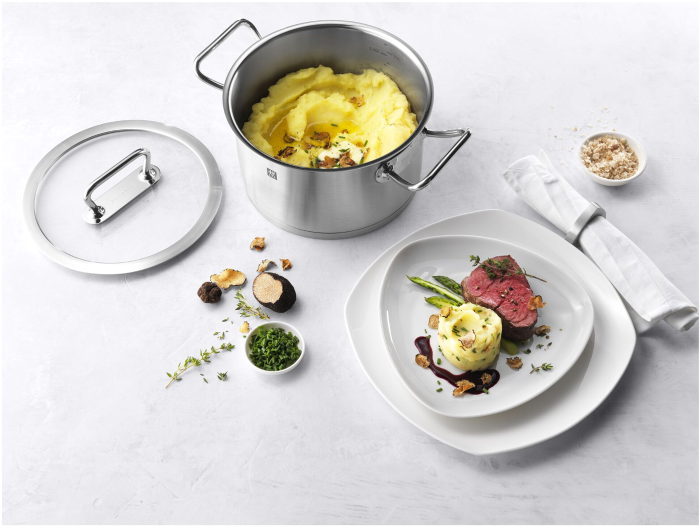 Ein Topf mit Kartoffelpüree, Teller mit Steak, Spargel und Kartoffeln. Trüffel und Kräuter sind auf einer weißen Oberfläche verstreut.