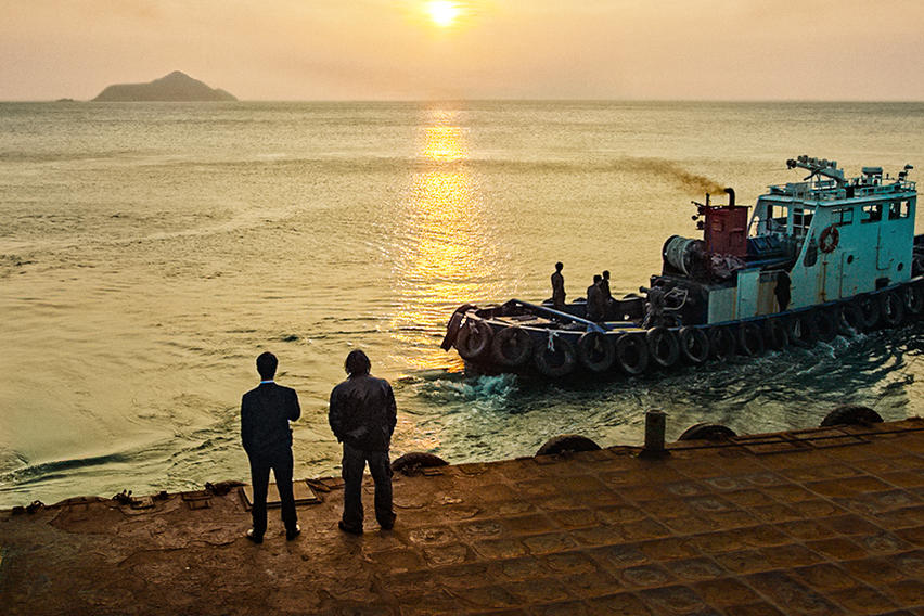 Zwei Männer auf einem Pier beobachten ein Boot mit Leuten, die auf dem Meer segeln. Die Sonne geht im Hintergrund unter.