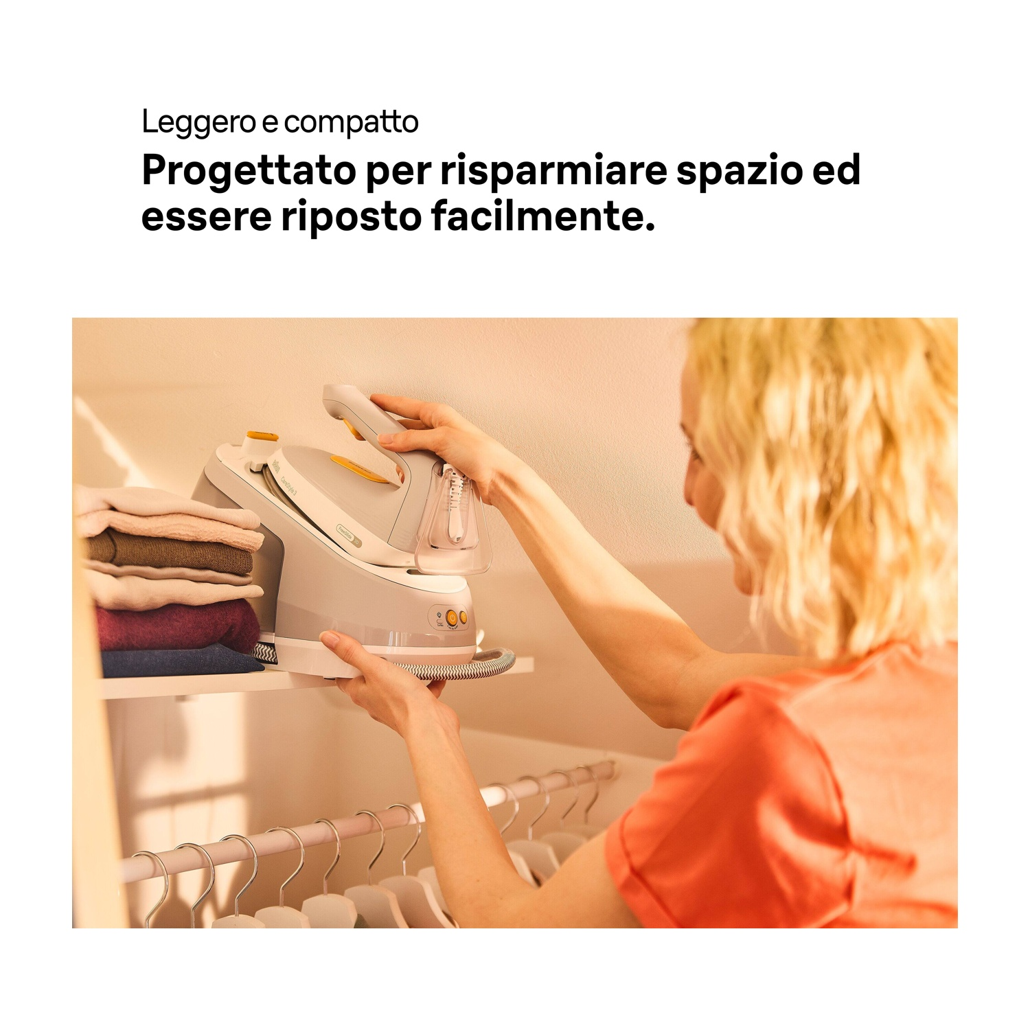 Una donna ripone un vaporizzatore per indumenti bianco e grigio su una mensola in un armadio. Gli abiti sono impilati sulla mensola. Sotto ci sono appendiabiti.