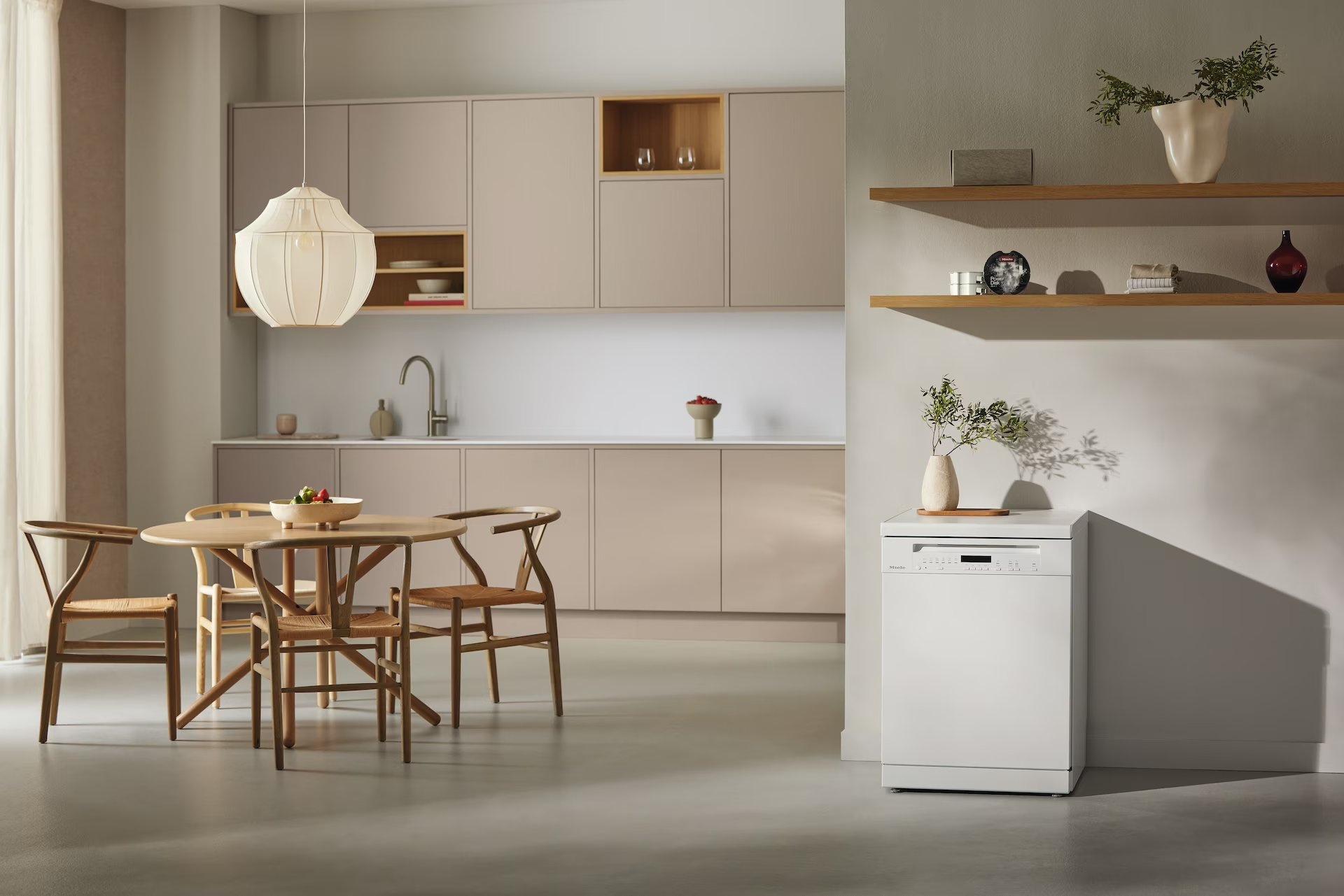 Una cocina con mesa y sillas, un lavavajillas blanco y armarios beiges. Hay una lámpara colgante sobre la mesa.