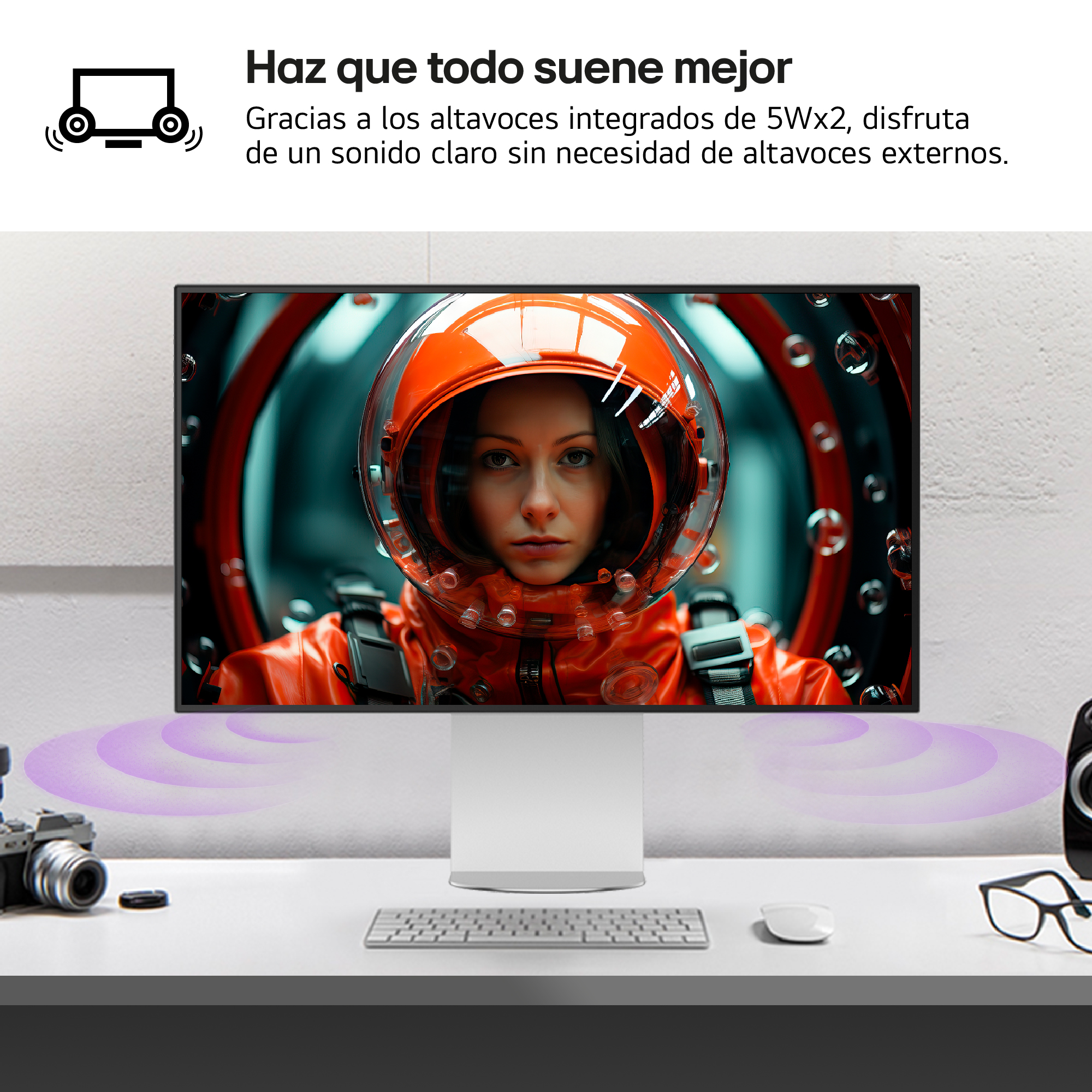 Una mujer con un traje espacial naranja está en un monitor. Un teclado, un ratón, gafas y una cámara están en el escritorio.