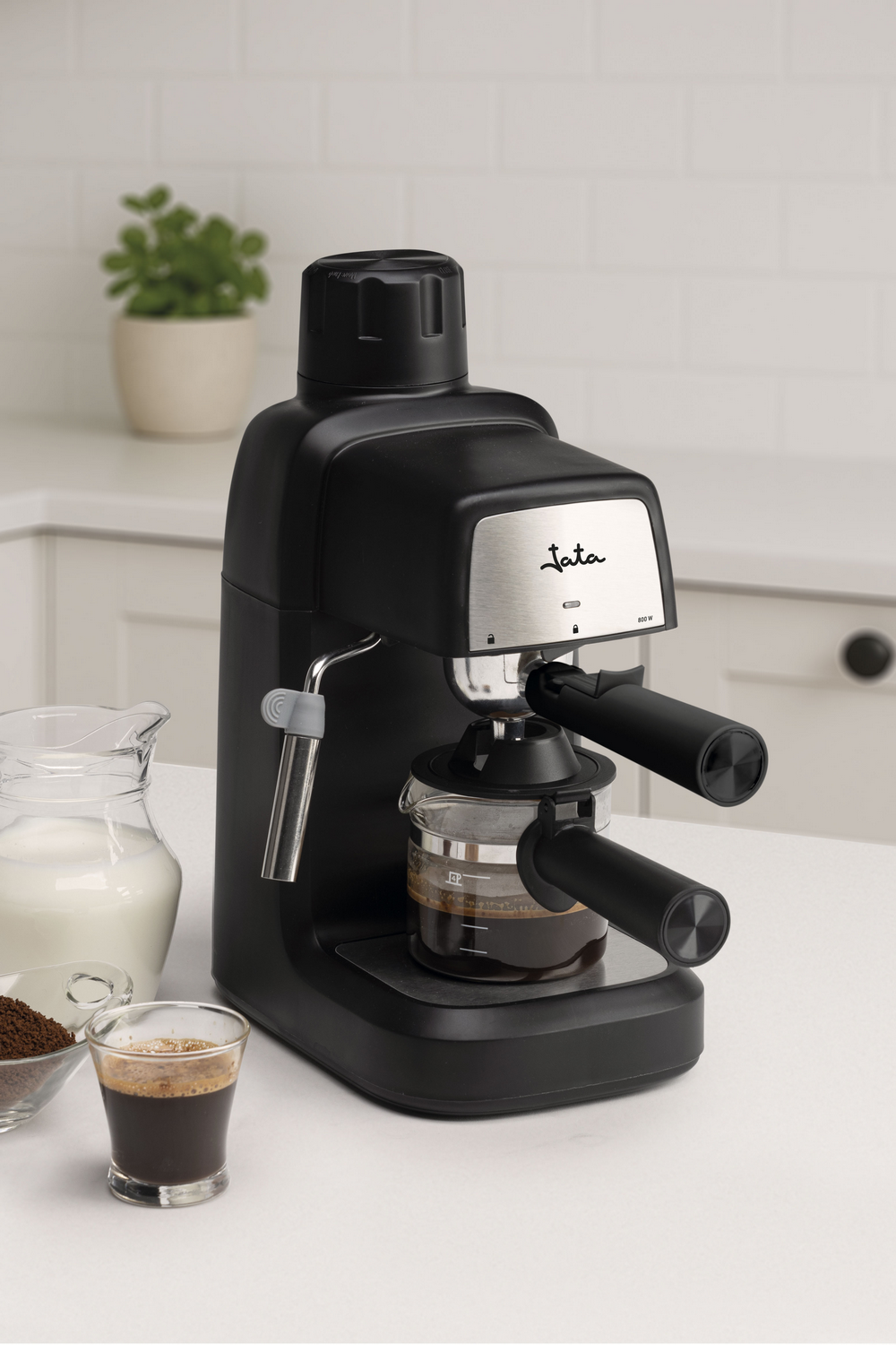 Máquina de espresso negra con café en vaso, leche, café molido y una pequeña planta sobre una encimera blanca.