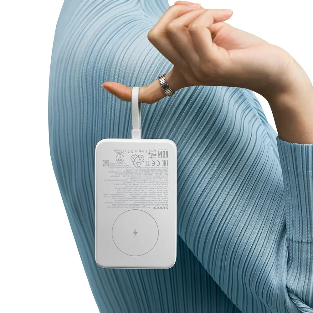 Una persona sostiene un power bank blanco con una mano. La persona lleva un anillo y una camisa plisada azul claro. El fondo es blanco.