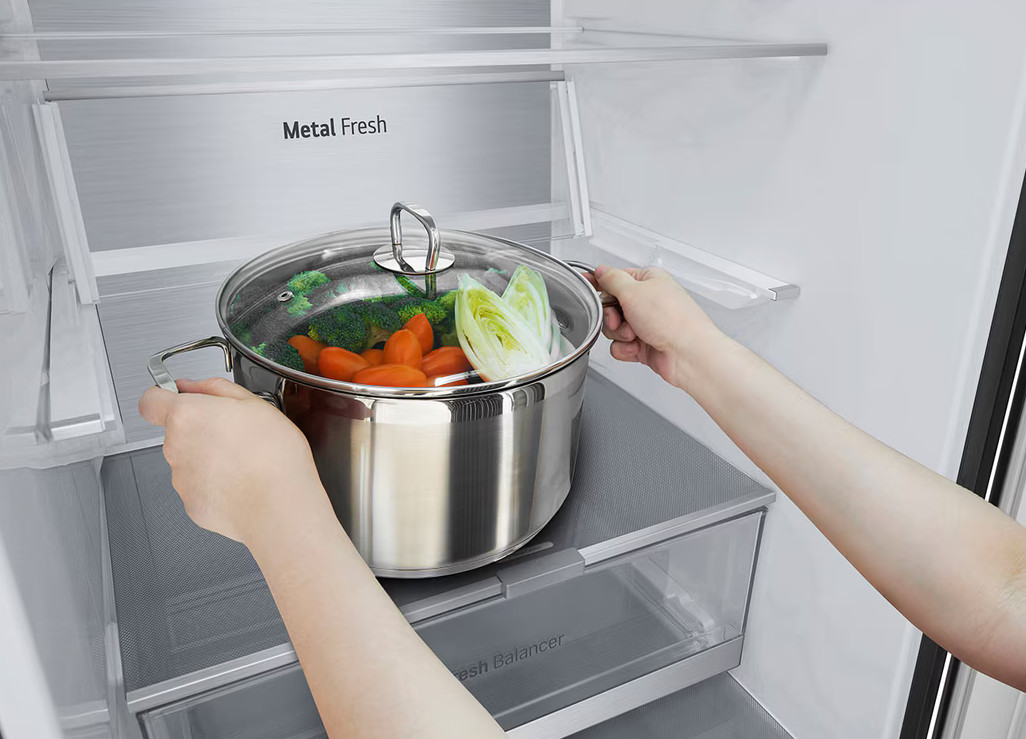Ręce wkładają garnek z warzywami do lodówki. Garnek zawiera marchewkę, brokuły i sałatę.