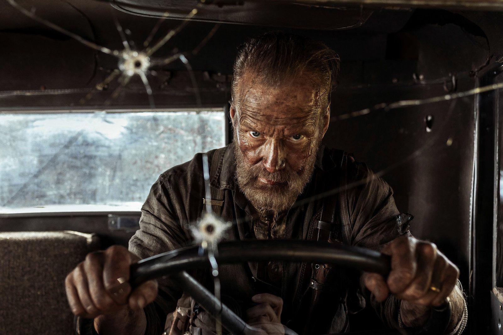 Ein Mann mit Bart in einem Auto blickt in die Kamera. Die Windschutzscheibe hat ein Einschussloch. Er hält das Lenkrad.