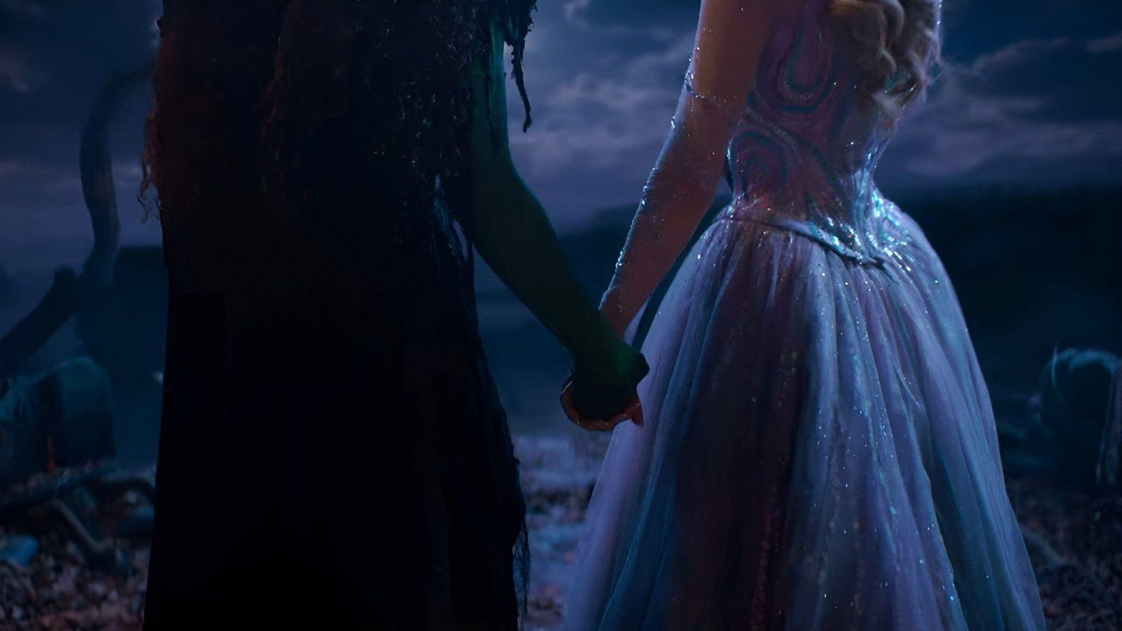 Zwei Frauen halten Händchen, eine in grünem Kleid und die andere in hellblauem Kleid, in dunkler Umgebung.