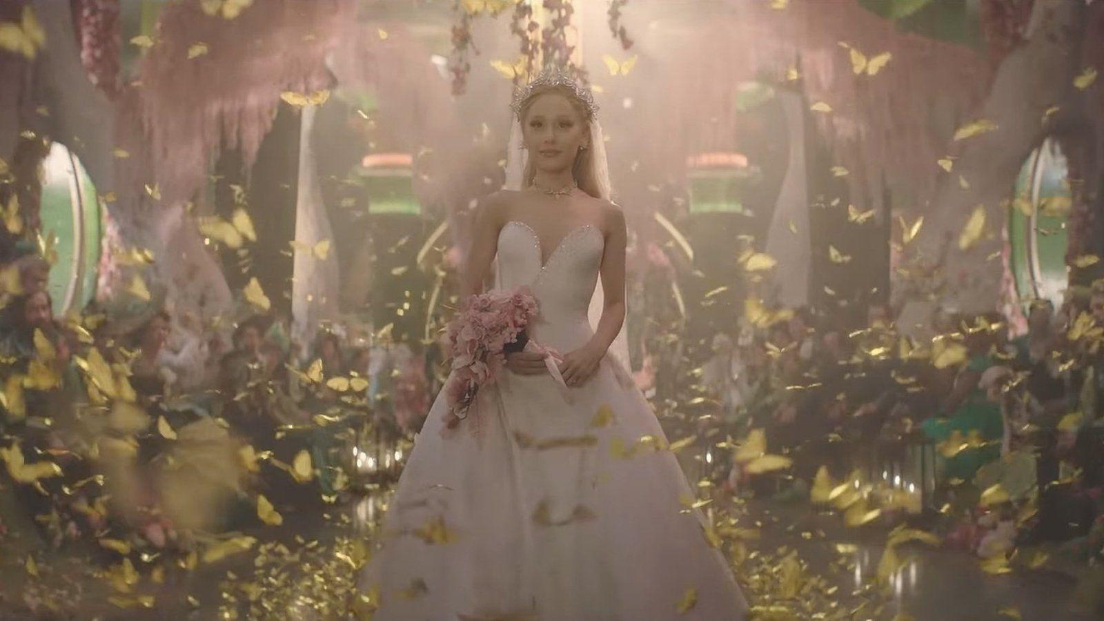 Eine Frau in weißem Kleid und Tiara hält Blumen. Gelbe Schmetterlinge schweben in einer rosa Halle mit Publikum.