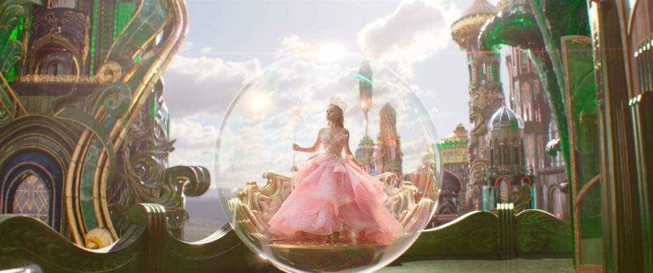Eine Frau in rosa Kleid und Diadem steht in einer Blase. Aufwendige Gebäude und ein sonniger Himmel bilden den Hintergrund.
