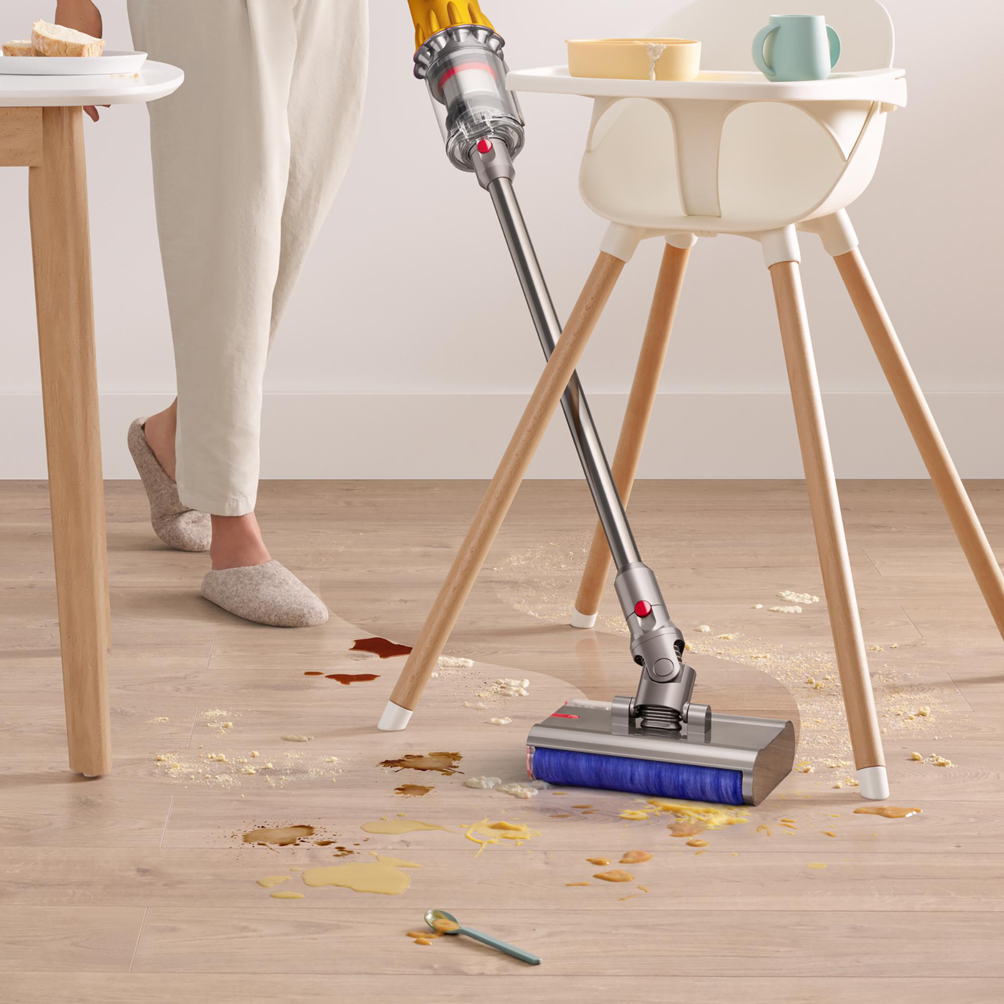Eine Person saugt einen unordentlichen Boden mit einem Staubsauger. Ein Hochstuhl und ein Tisch sind im Hintergrund.