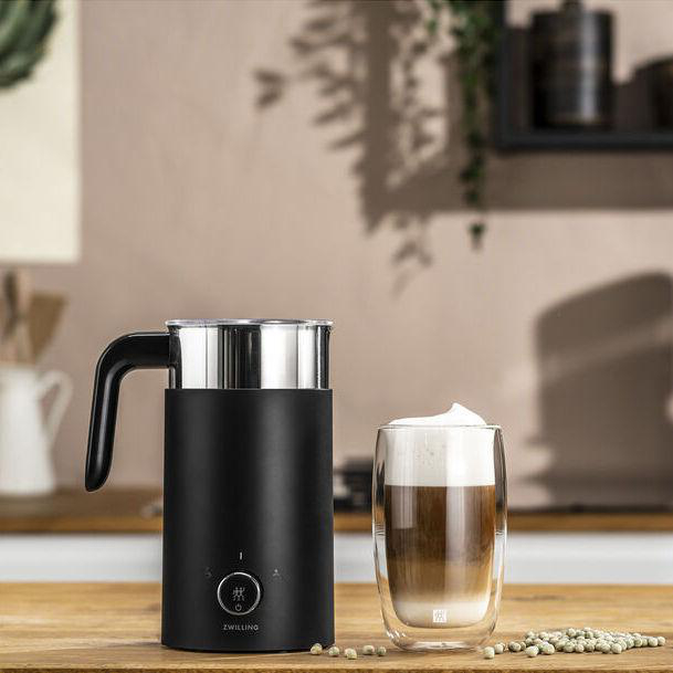 Ein schwarzer Milchaufschäumer und ein Glas Latte auf einer Holzoberfläche. Grüne Samen sind verstreut. Hintergrund ist eine Küche.
